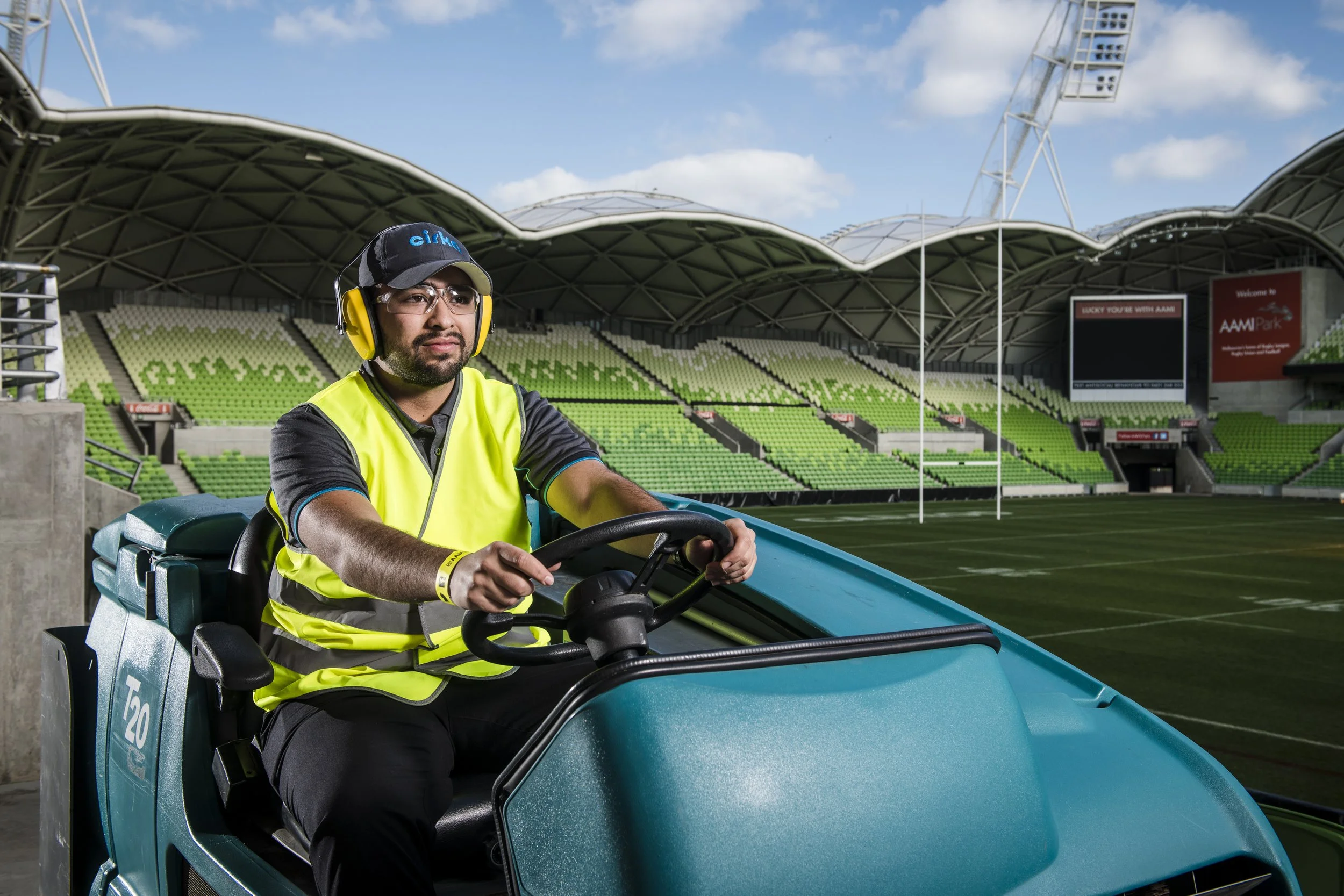 A man wearing a yellow safety vest, glasses, a cap, and ear protection driving a small blue vehicle on a football stadium field.