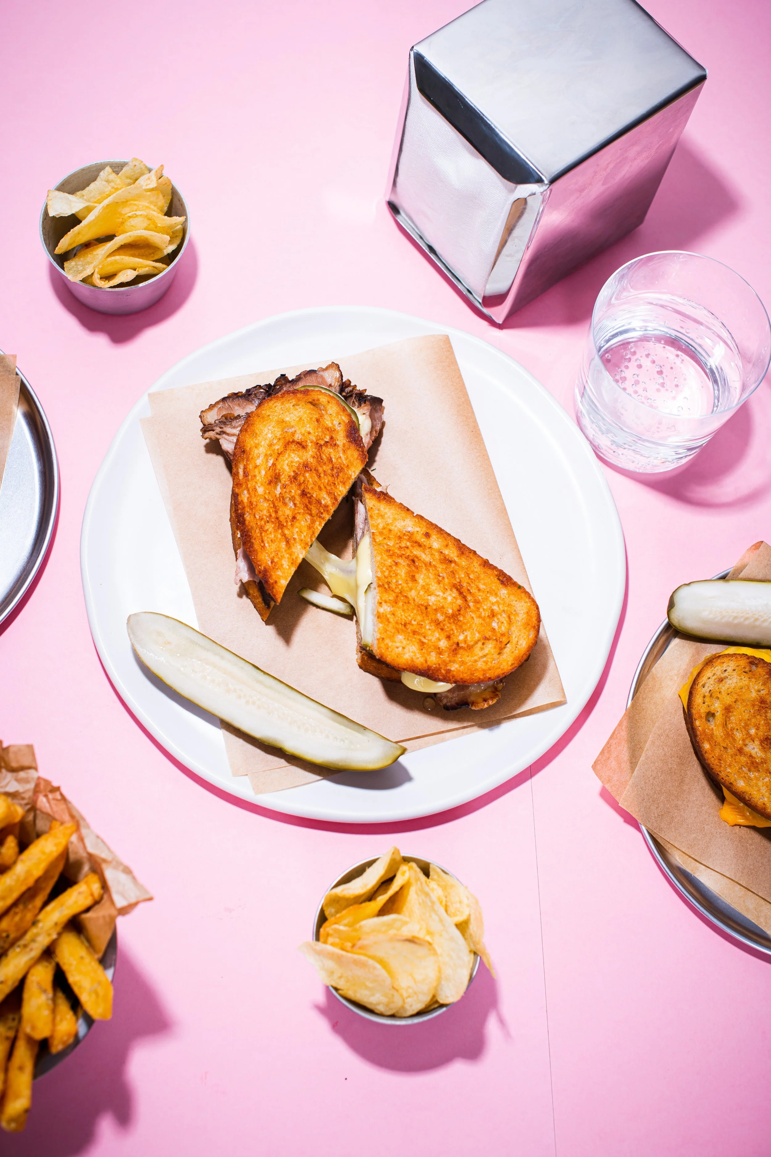 A pink table with a plate of fried chicken sandwich, pickle slices, a cup of potato chips, a glass of water, and small bowls of potato chips and French fries.