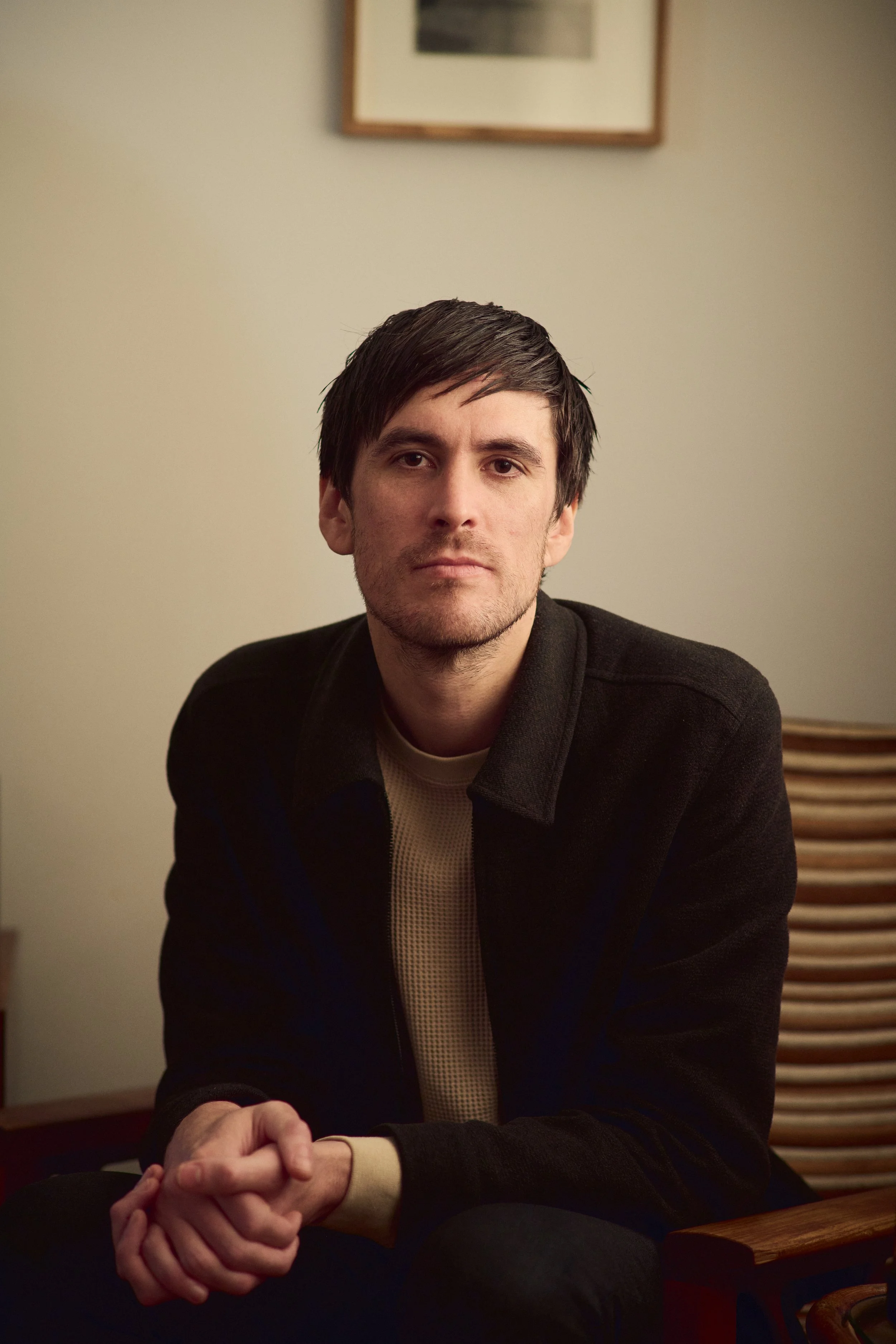 A young man with dark hair and light skin sitting at a table with hands clasped.