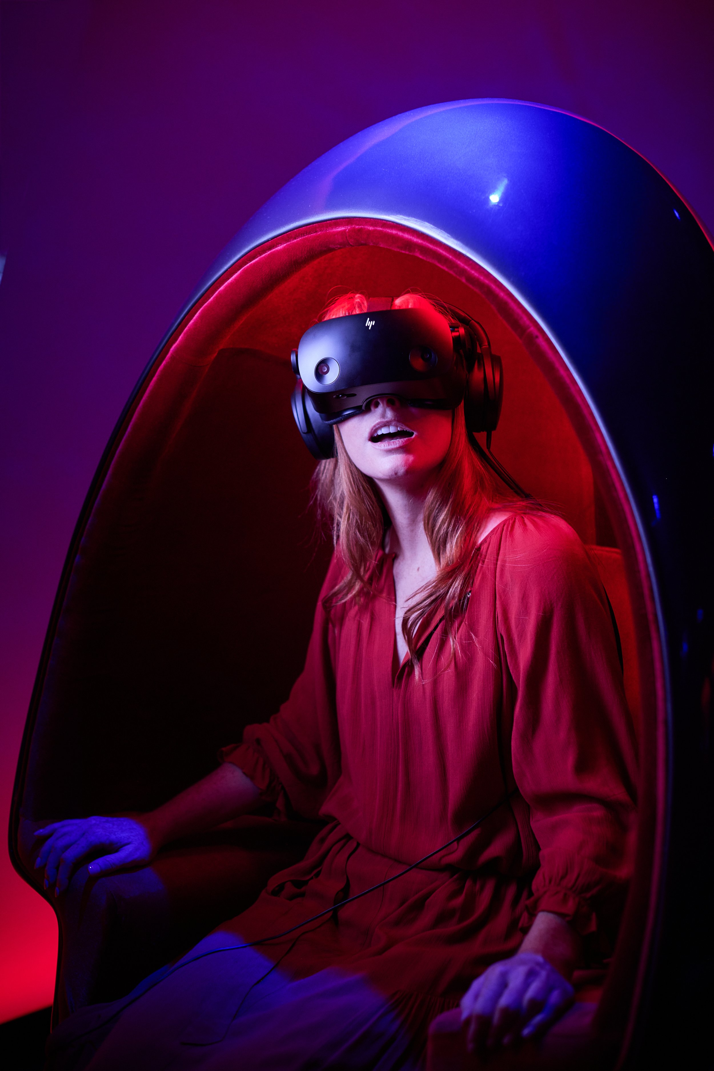 A woman with red hair seated inside a pod with rounded, illuminated walls, wearing a virtual reality headset, in a colorful, neon-lit environment.