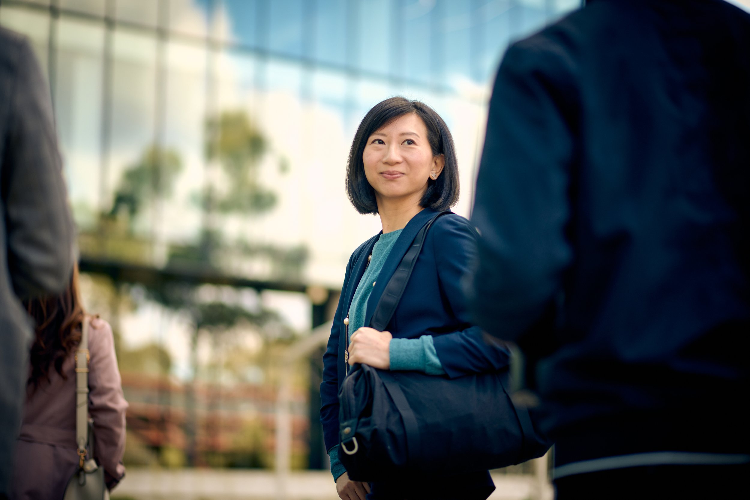 A woman with shoulder-length black hair smiling, wearing a navy blazer and teal top, engaged in conversation with individuals around her in an outdoor urban setting.