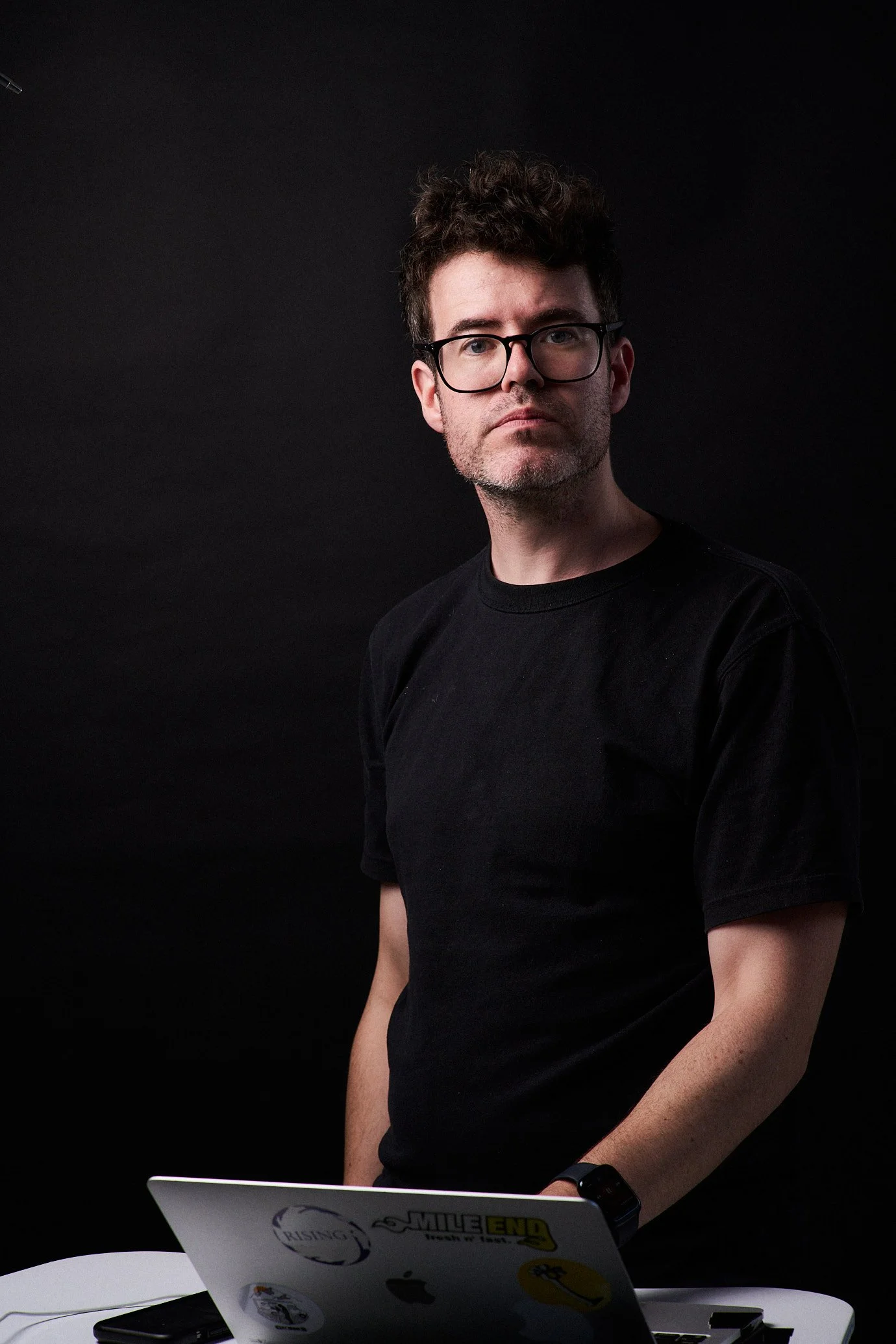 A man with curly dark hair, glasses, and a slight beard wearing a black T-shirt, standing behind a white table with a laptop, against a black background.
