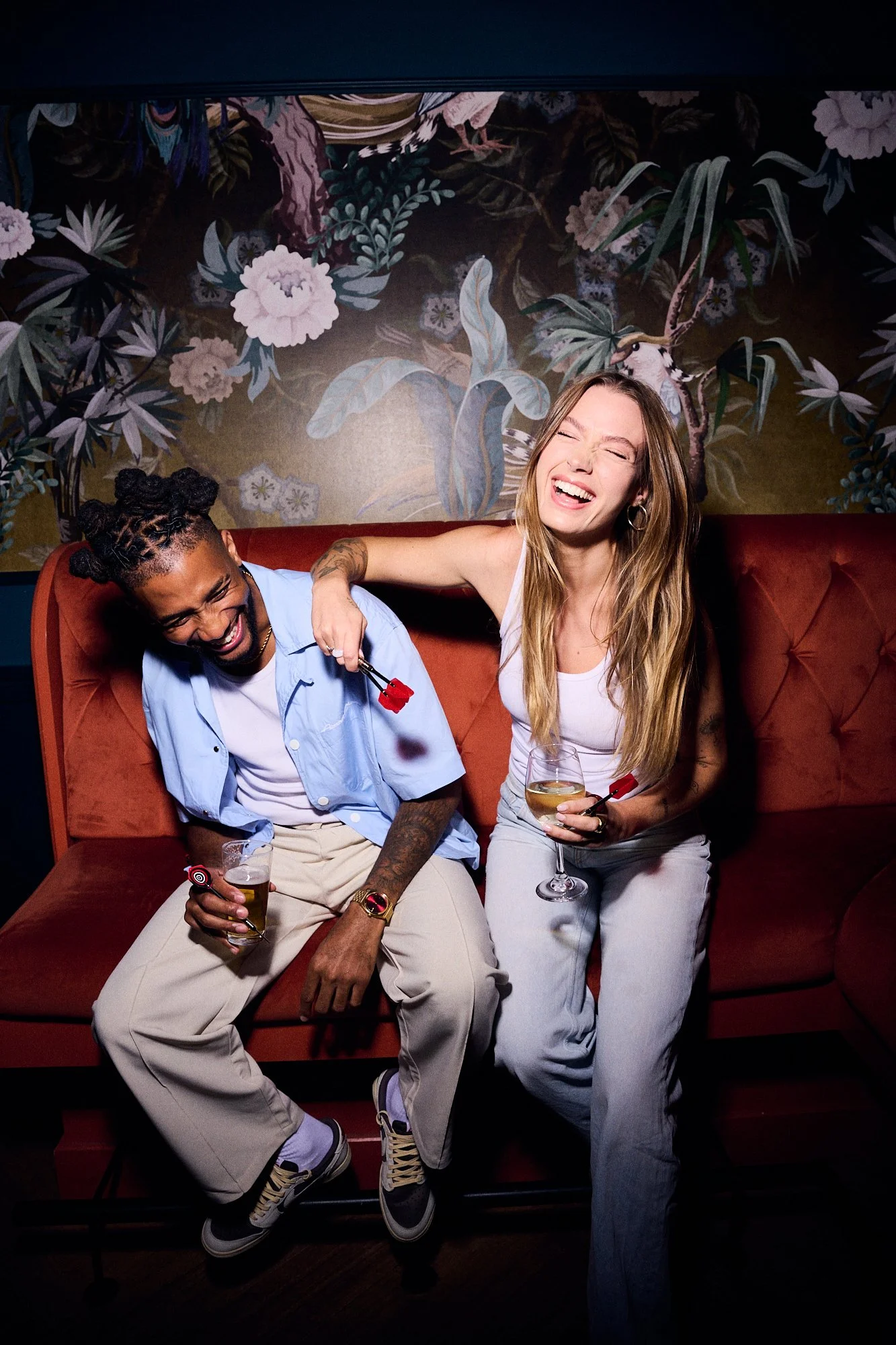 Two friends sitting on a red velvet sofa, laughing and enjoying drinks at a bar or lounge with a lush jungle-themed wall in the background. The person on the left is a man with braids wearing a blue shirt and beige pants, holding a drink, while the w