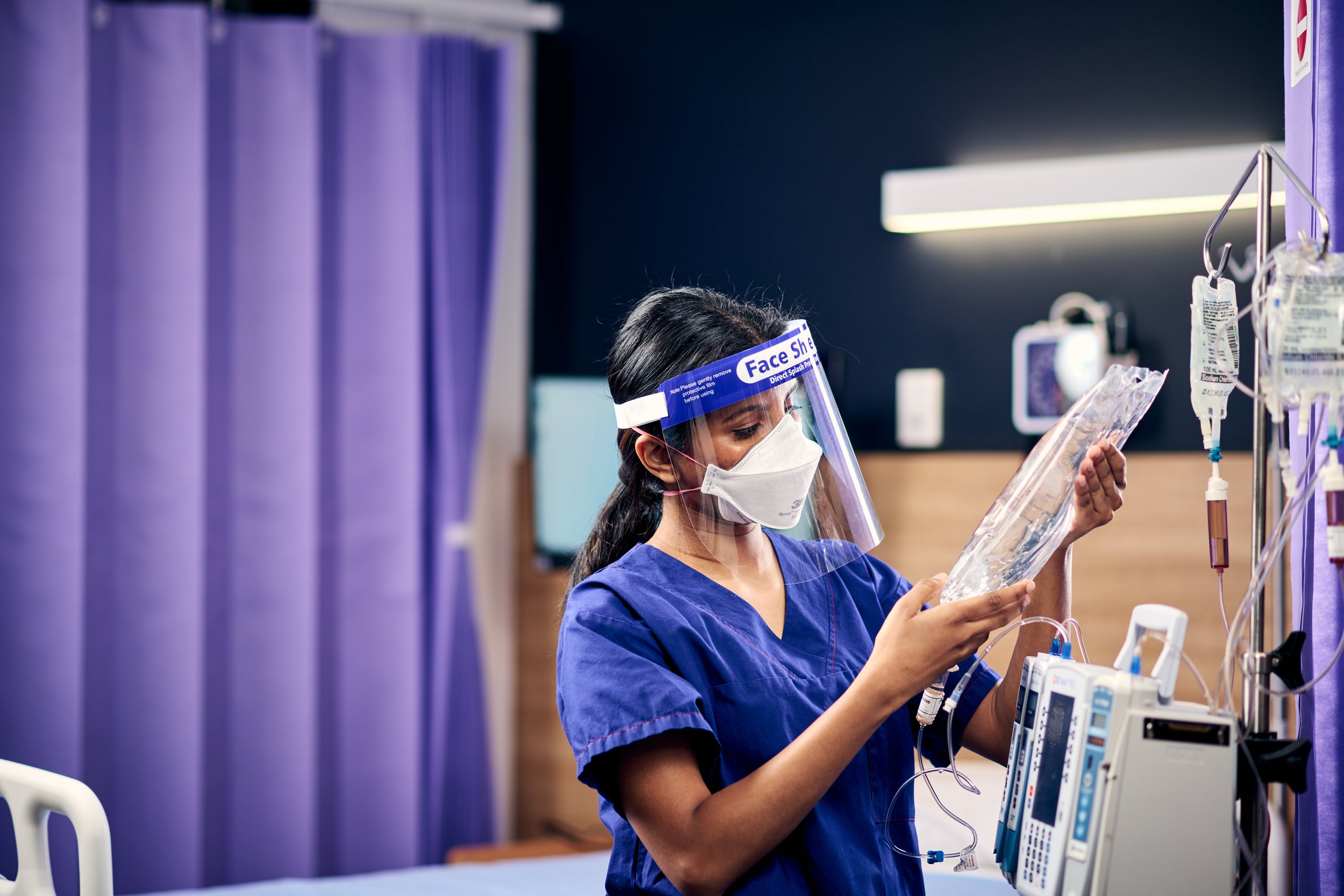 A healthcare worker wearing a face mask, face shield, and blue scrubs preparing medical equipment in a hospital setting.