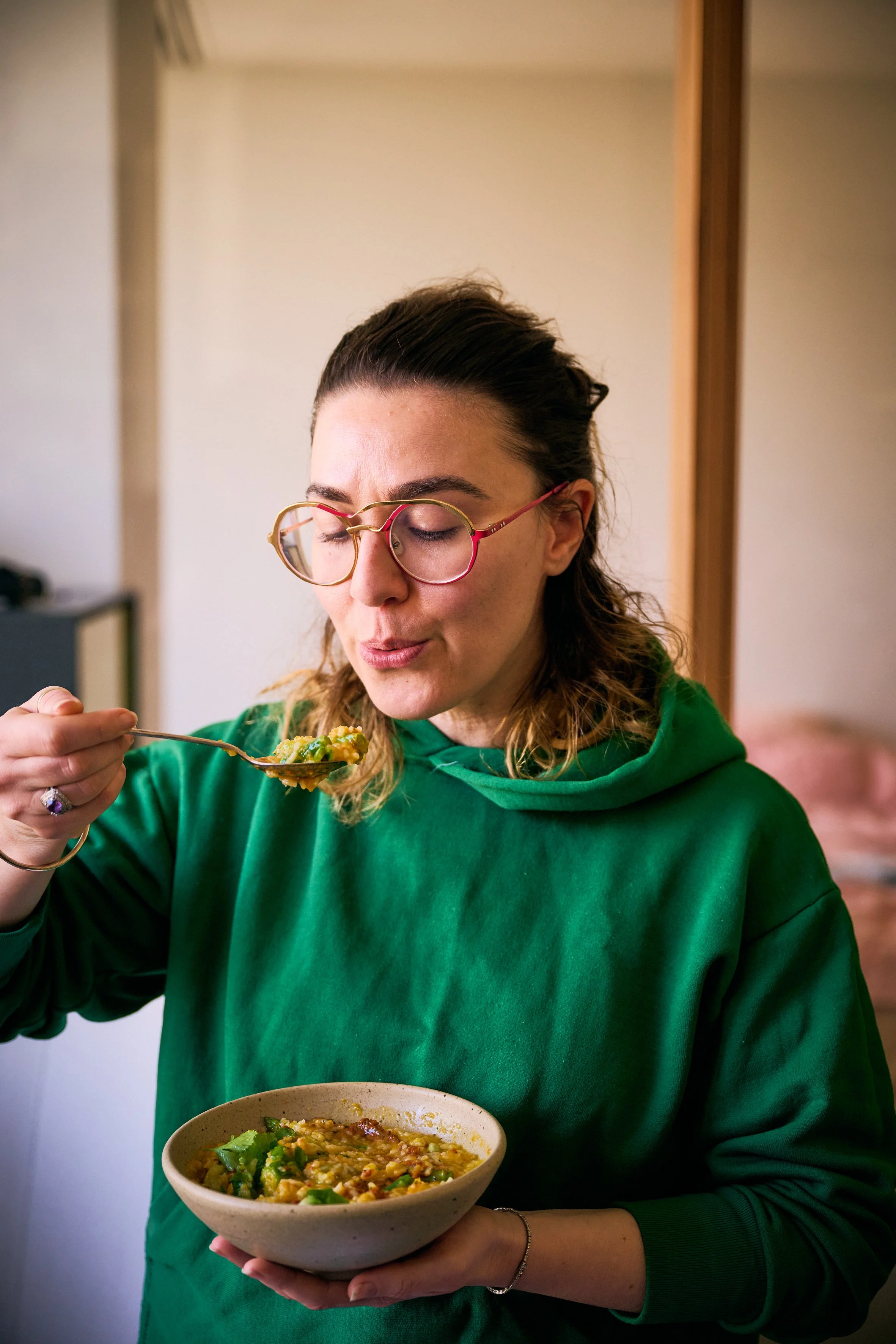 A woman wearing a green hoodie and glasses is sitting indoors, holding a beige bowl of food in her left hand and using a spoon to eat with her right hand. The food appears to be a dish with rice, vegetables, and possibly some meat.