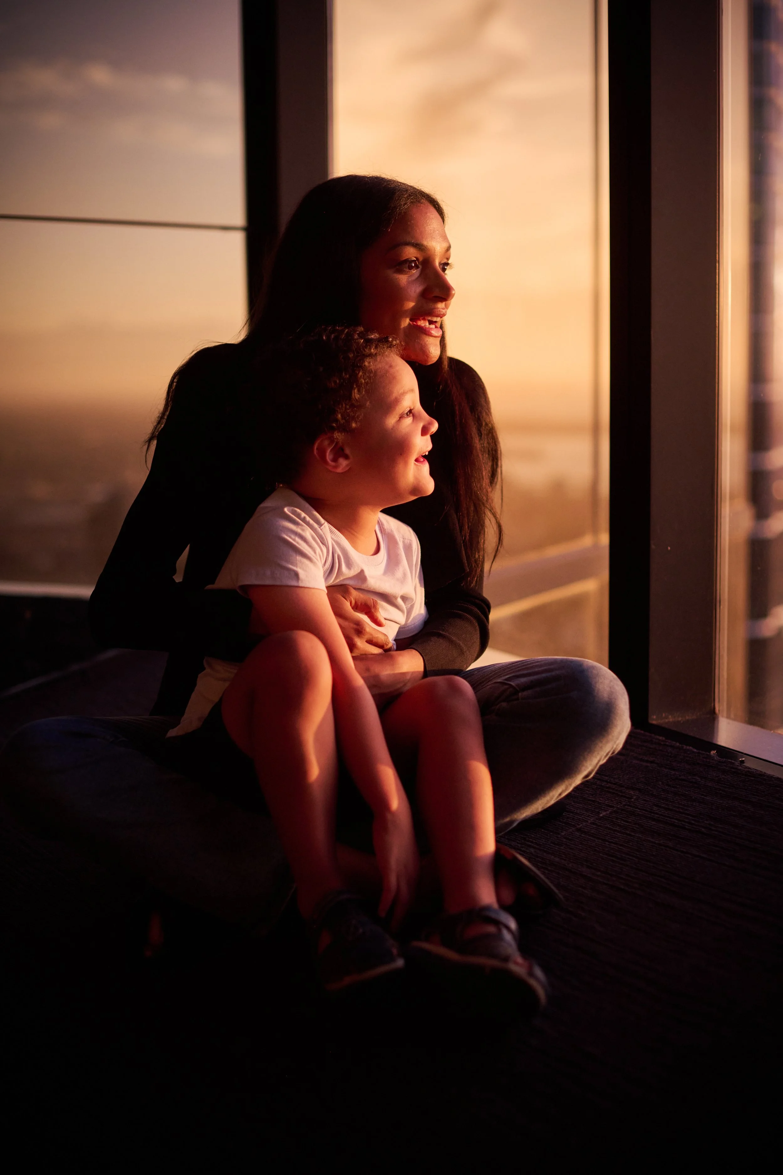 A woman and a young girl sitting together on a mat, looking out a large window at sunset. They are smiling and enjoying the view.