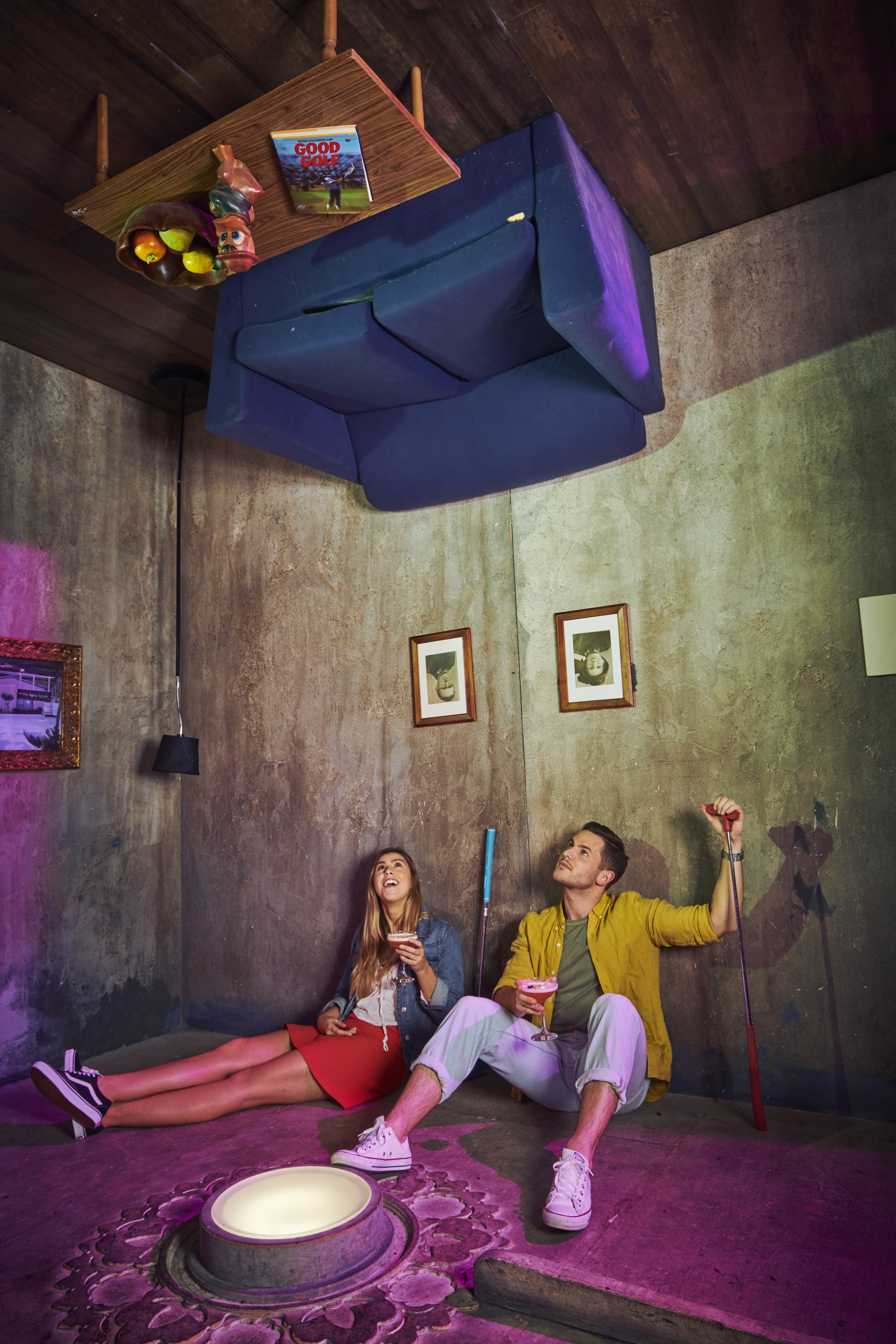 A man and woman sitting on the floor in a room with dark walls, holding drinks and looking up at a table that is floating upside down on the ceiling. The table has a fruit bowl and a magazine on top. They appear to be enjoying themselves in a playful