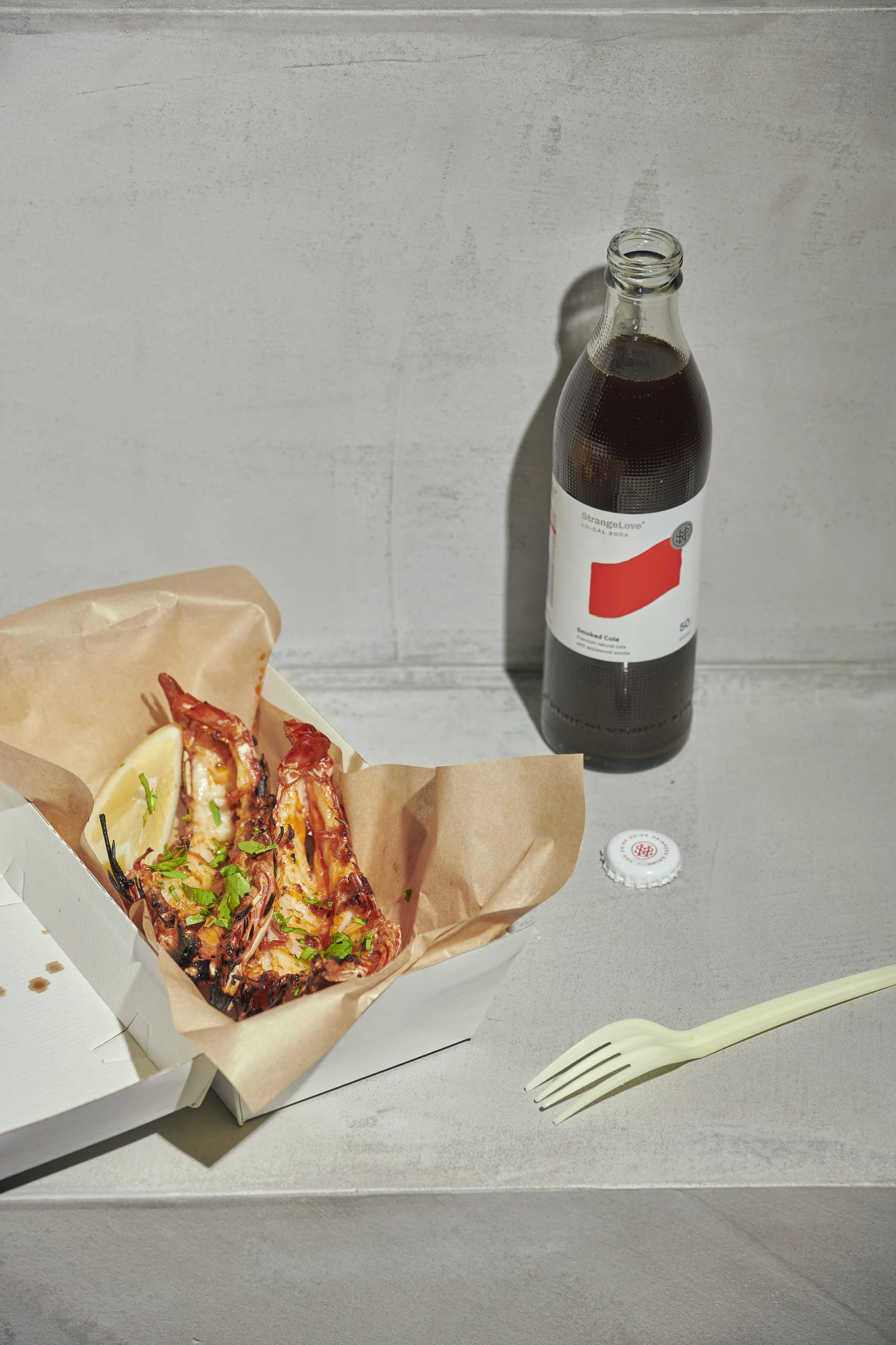 Lobster roll in a paper-lined container next to a bottle of black soda on a gray table, with a fork nearby.