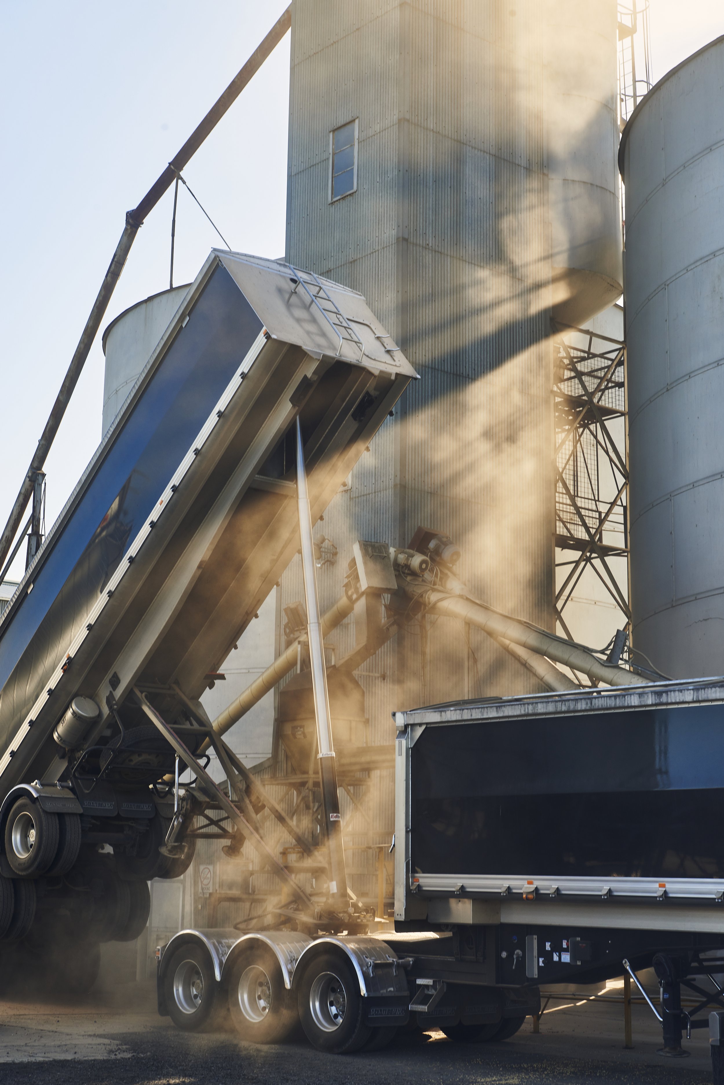 A semi-truck with an open trailer is unloading or loading steel pipes through a conveyor system, with dust and sunlight in the background.