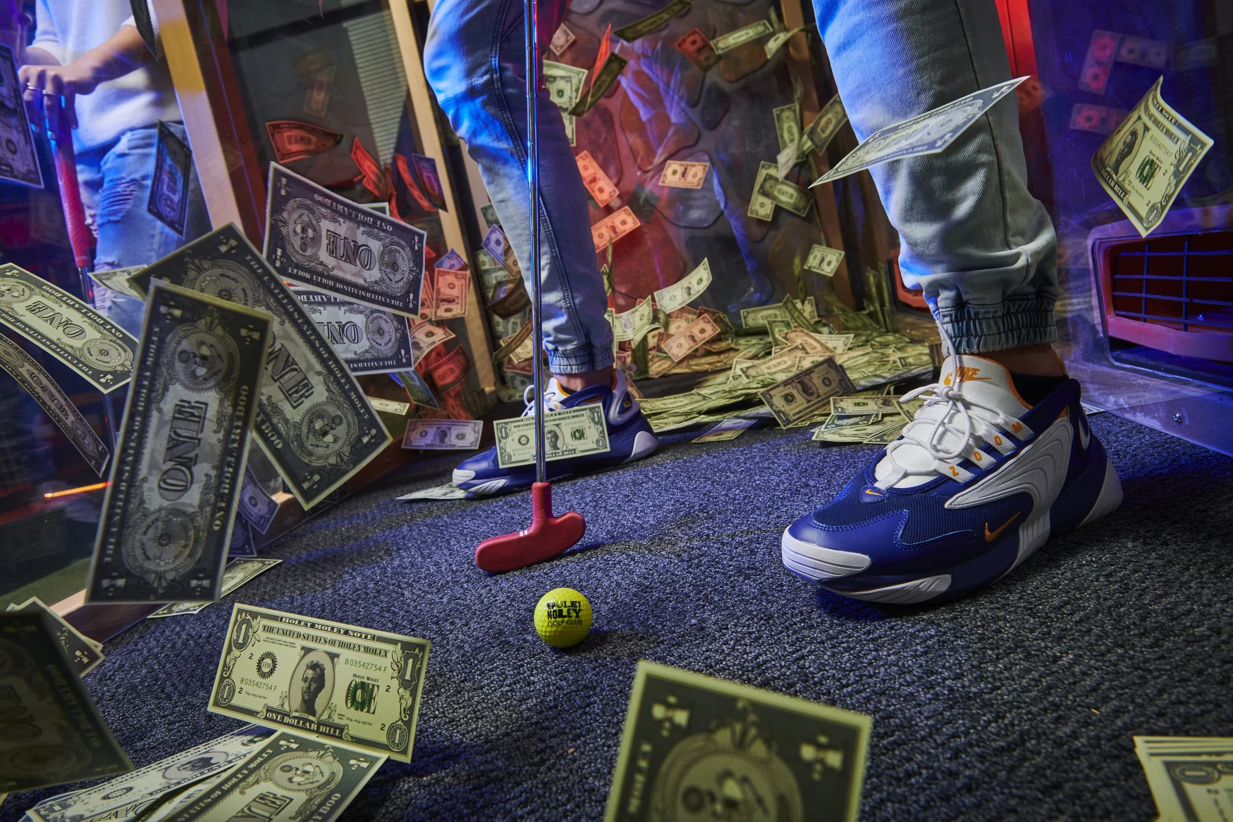 Someone standing inside a glass enclosure with an abundance of dollar bills scattered on the floor and floating in the air. The person is wearing jeans and sneakers, and a yellow golf ball with the text 'Holey Moley Golf Club' is on the ground.