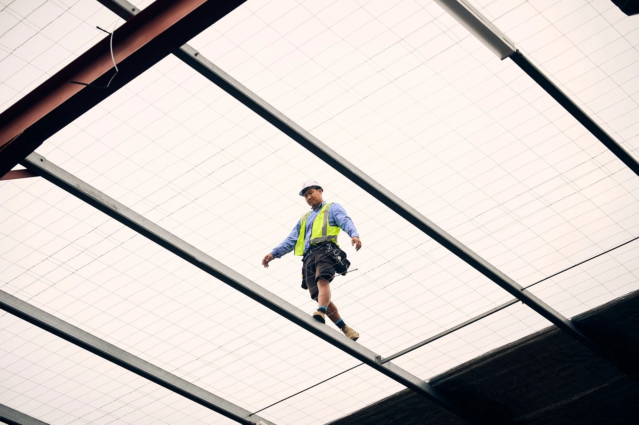 A worker wearing a safety helmet, safety vest, and work attire walking on a steel beam beneath a large, grid-patterned roof structure.