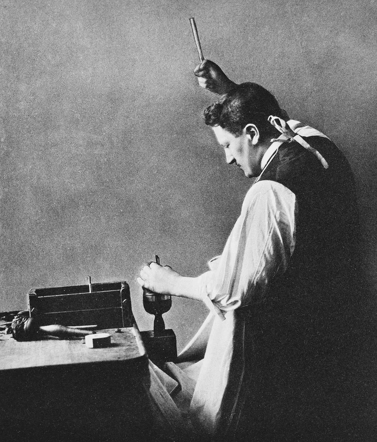 Photograph of Mr Joseph Asscher standing to the right of a bench while holding a cutting tool resting on the Cullinan Diamond, held in a vice. He holds a hammer in his raised right hand about to strike the cutting tool. Image: Royal Collection © Her Majesty Queen Elizabeth II
