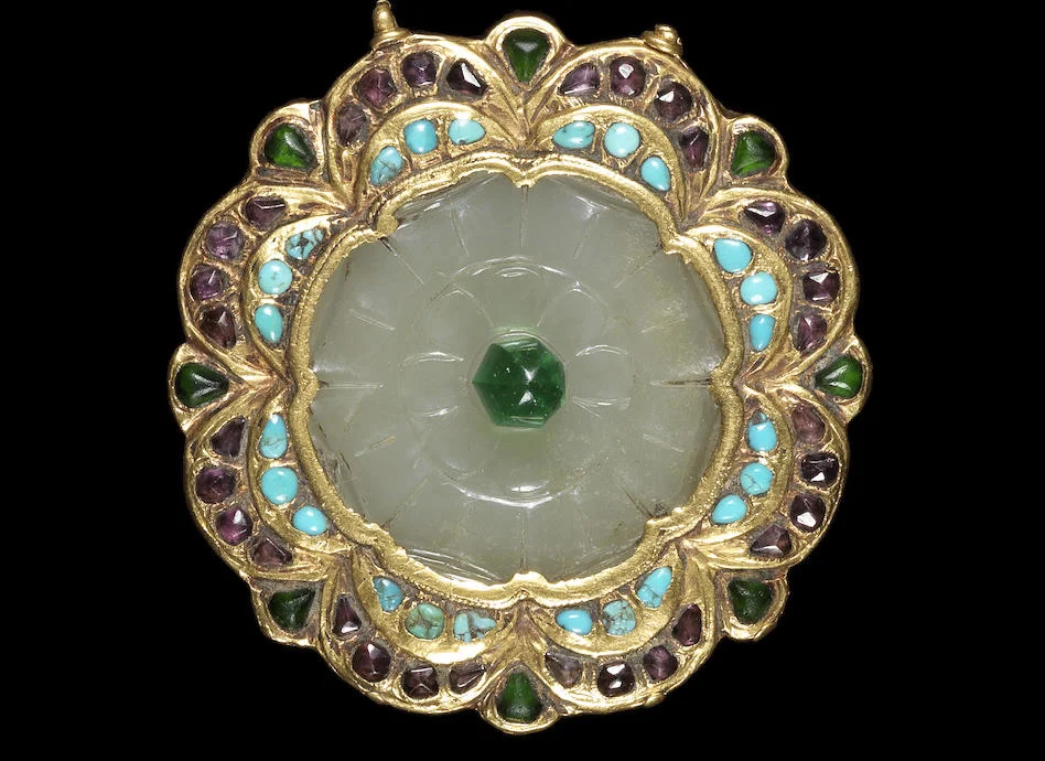 A Qajar gem-set and gold mounted jade Plaque. Persia, 19th Century. In the form of a flowerhead, the jade carved with petals and surmounted by a green glass bead, surrounded by a stepped gold mount set with turquoise and paste, small gold attachment loop to side and two jade attachment loops to reverse. 5.8 cm. diameter. Image: Bonhams
