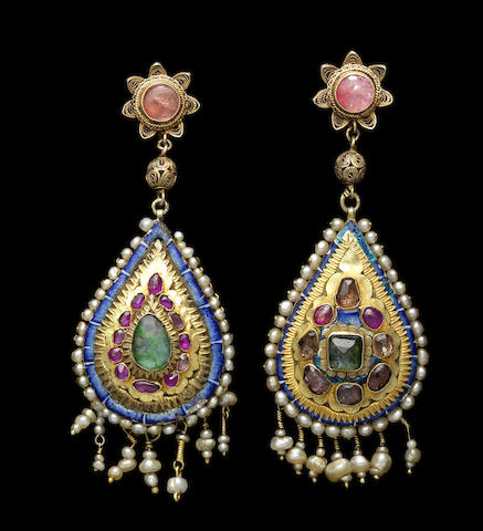 Two Qajar gem-set gold Earrings. Persia, 19th Century. Each of drop-shaped form, with gem-stones in raised gold settings, surrounded by a narrow blue enamelled border and a seed pearl edge, with seed pearl suspensions on twisted gold wire, verso with polychrome floral enamelled decoration, surmounted by gem-set floral and globular gilded silver filigree elements probably added later, with screw and loop. Image: Bonhams