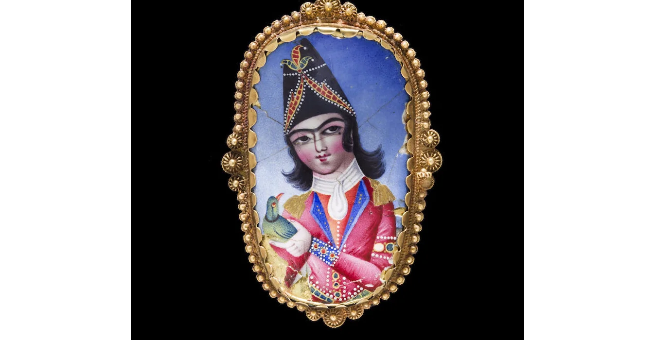 A Qajar enamelled plaque mounted as a gold brooch. Persia, 19th Century. Of curved oval form, the polychrome enamel plaque depicting an elegantly dressed courtier holding a parakeet, the yellow gold mount decorated in filigree. Image: Bonhams