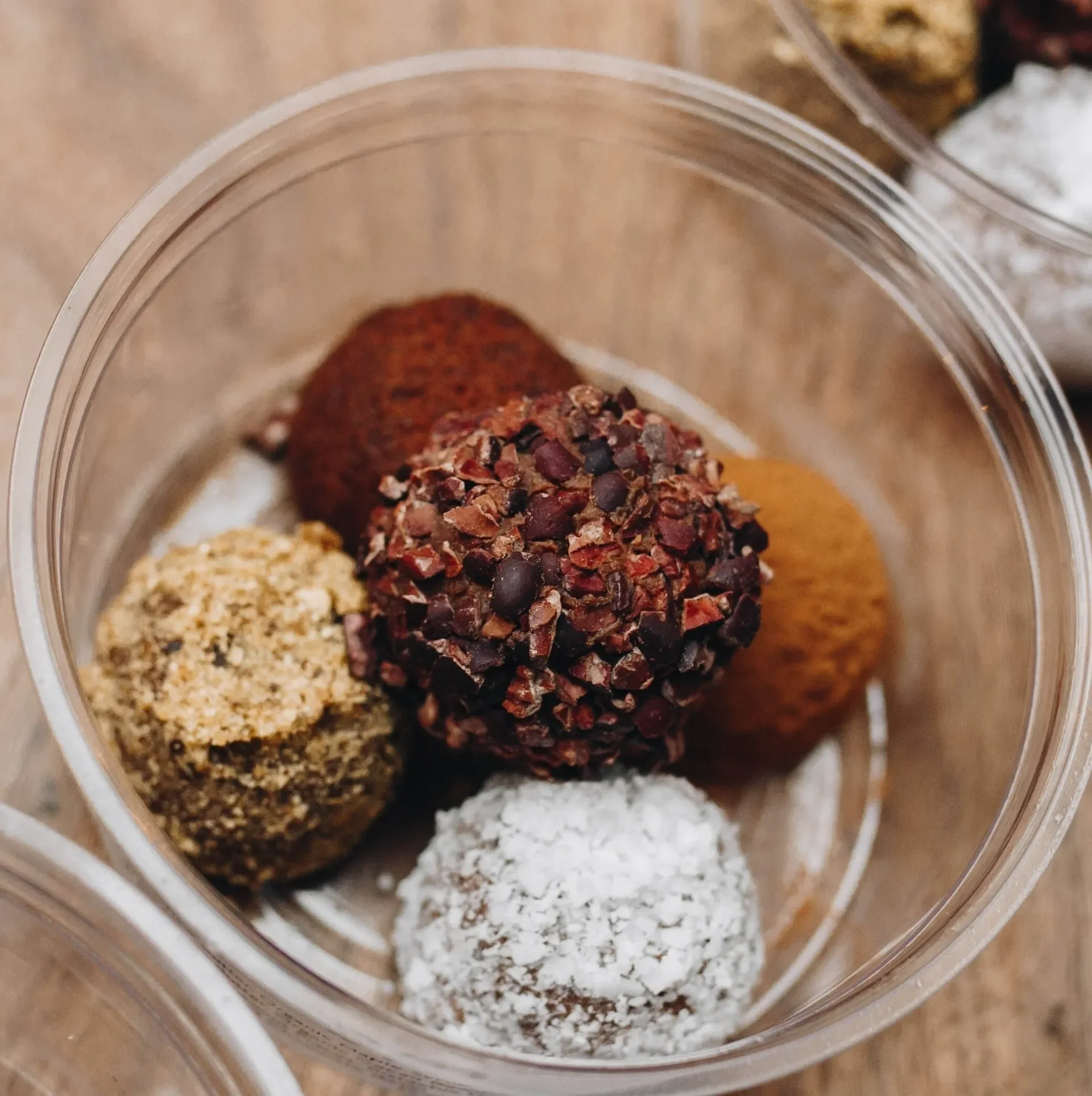 Assorted chocolate truffles in a clear container, coated with toppings like cacao nibs, shredded coconut, and crushed nuts.