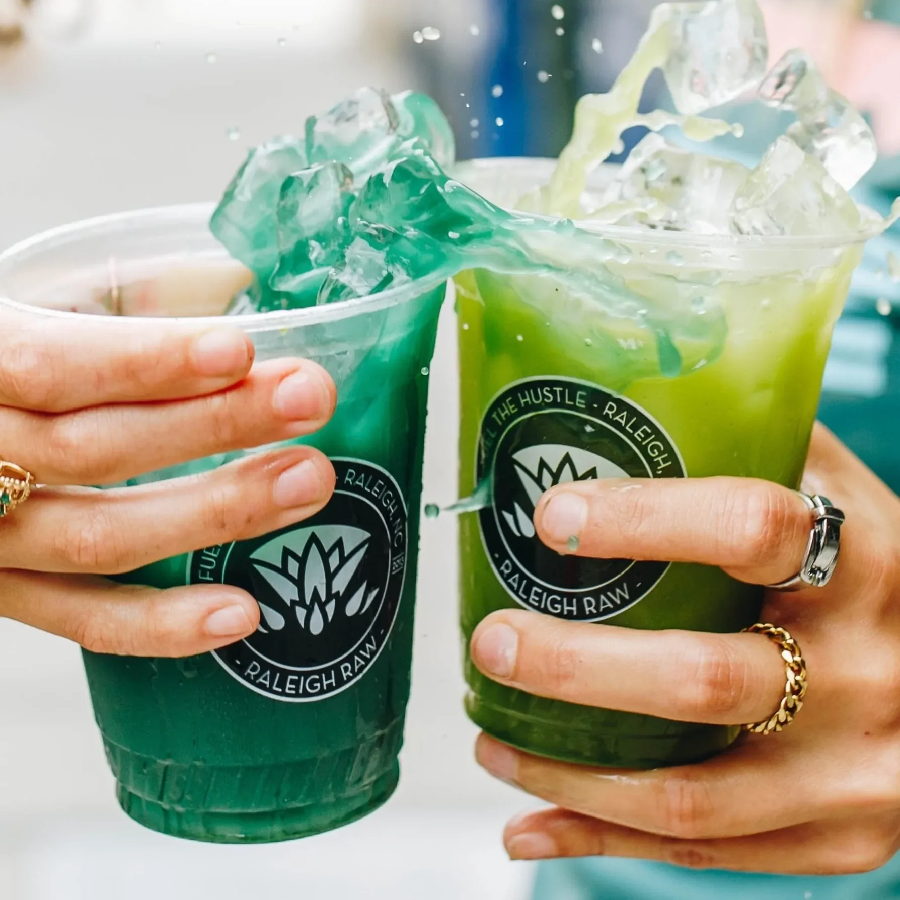 Vibrant Raleigh Raw matcha lemonade and blue spirulina lemonade clinking together in a refreshing splash. These energizing, antioxidant-rich drinks offer bright, clean hydration with superfood ingredients and bold summer flavor.