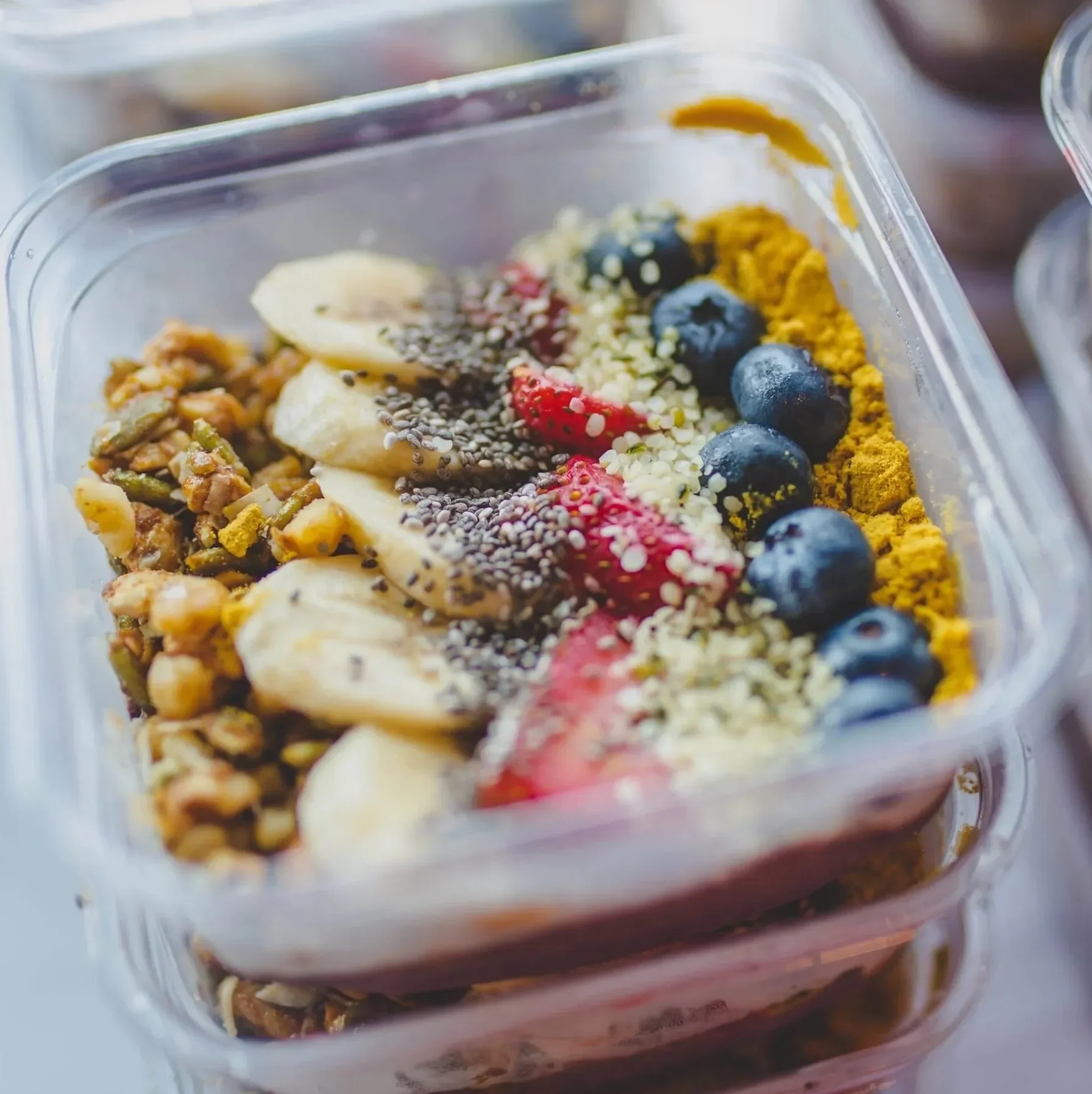 Close-up of a layered parfait in a clear to-go container topped with banana slices, strawberries, blueberries, granola, chia seeds, hemp seeds, and turmeric powder