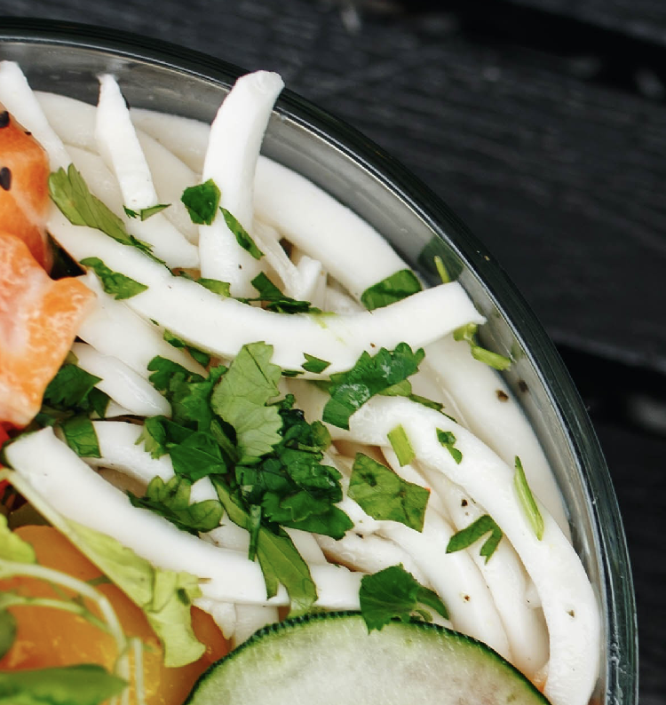 Close-up of a fresh coconut ceviche bowl featuring sliced white coconut strips topped with chopped cilantro, next to cucumber and colorful vegetables.