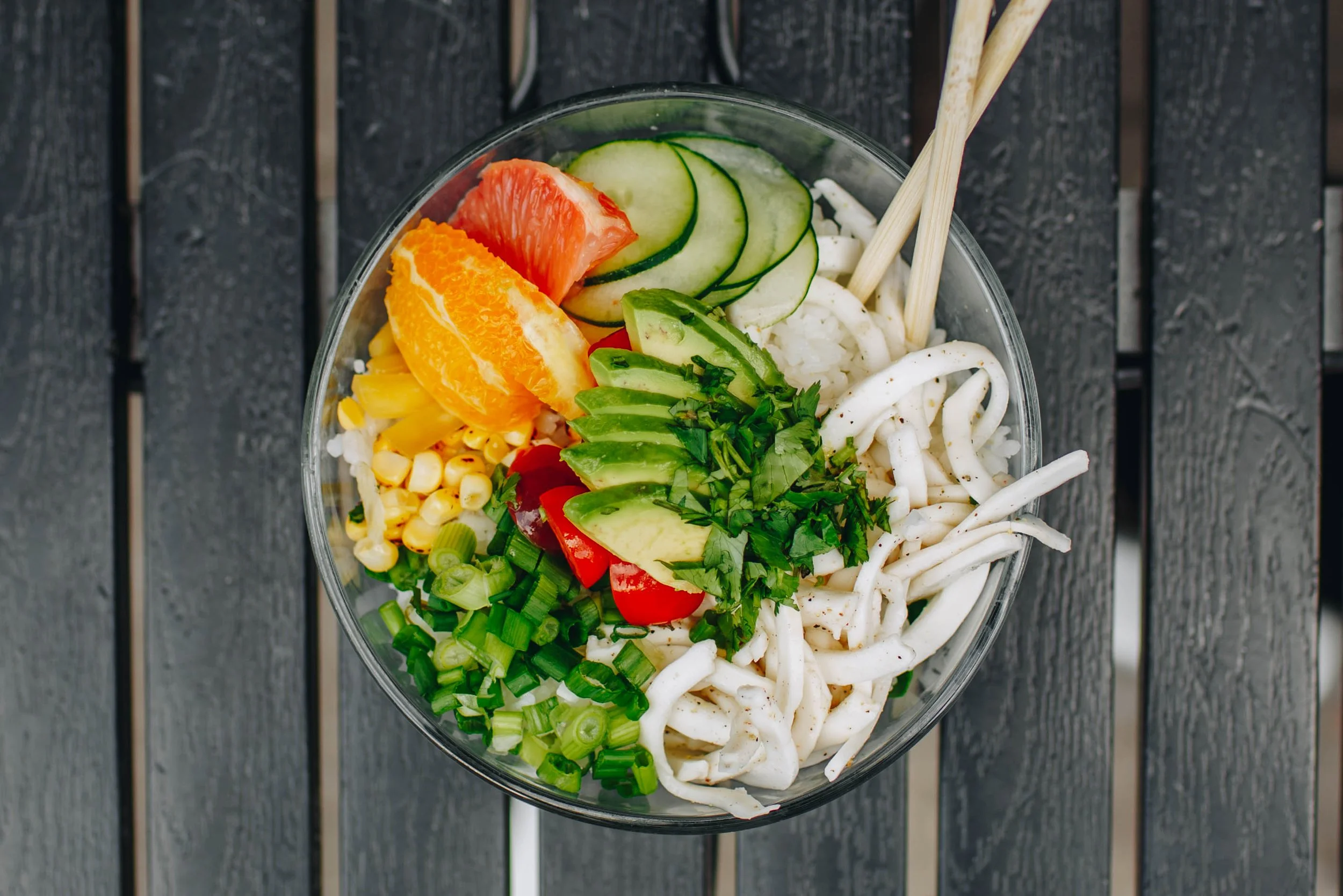 A colorful poke filled with sliced avocado, cucumber, citrus segments, corn, scallions, tomatoes, cilantro, and seasoned coconut ceviche arranged over rice, displayed on a dark wooden surface with chopsticks resting on the bowl.