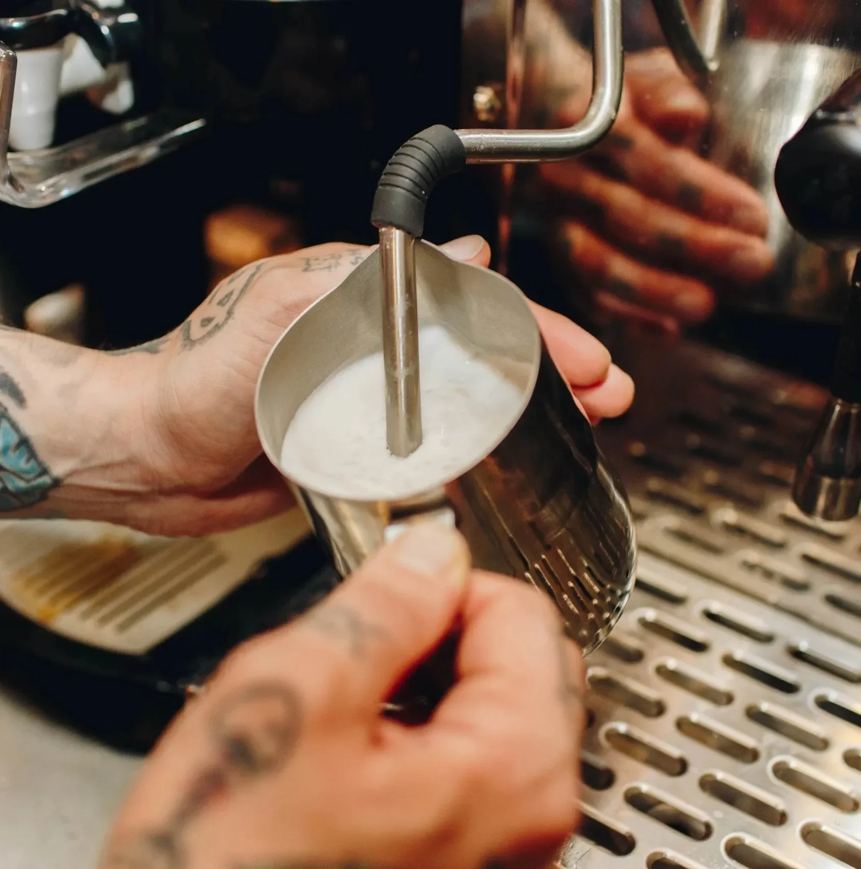 Close-up of a barista steaming milk to create silky, microfoam for a cappuccino. Stainless steel pitcher, skilled hands, and warm espresso machine details highlight Raleigh Raw’s craft-focused coffee preparation.