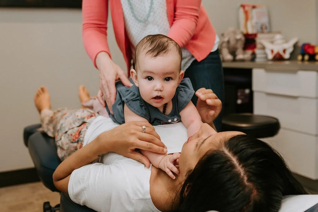 image description: a woman lays on her back on a chiropractor’s table with her infant laying on her chest. dr jessica de nooij, their chiropractor, gently adjusts the infant.