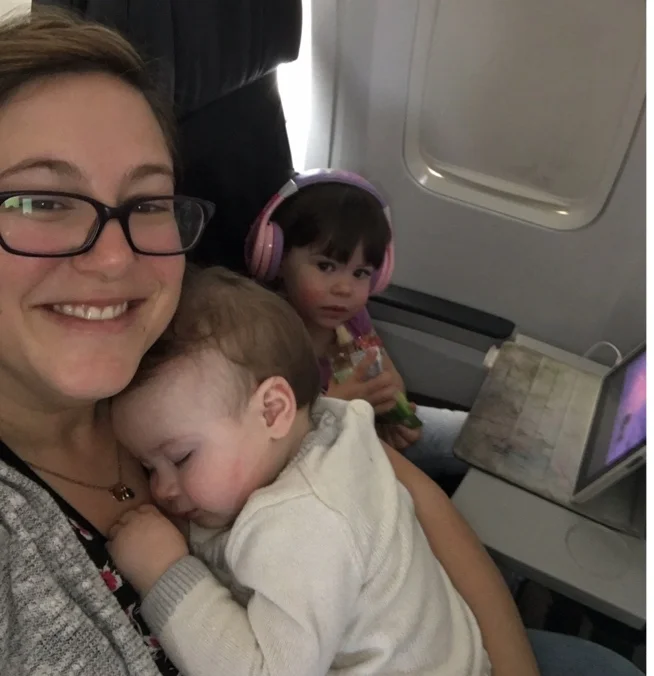 Image Description: Christine travels with her two young children, an infant sleeping on her chest and beside her a toddler watching a tablet with earphones holding a snack.