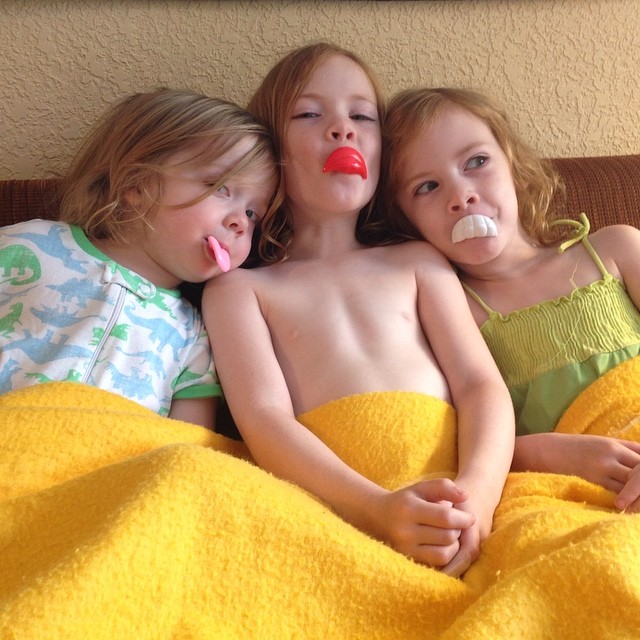Image Description: Rachel’s three kids sitting on a couch, snuggled under a yellow blanket, with Mr. Potato Head pieces in their mouths.