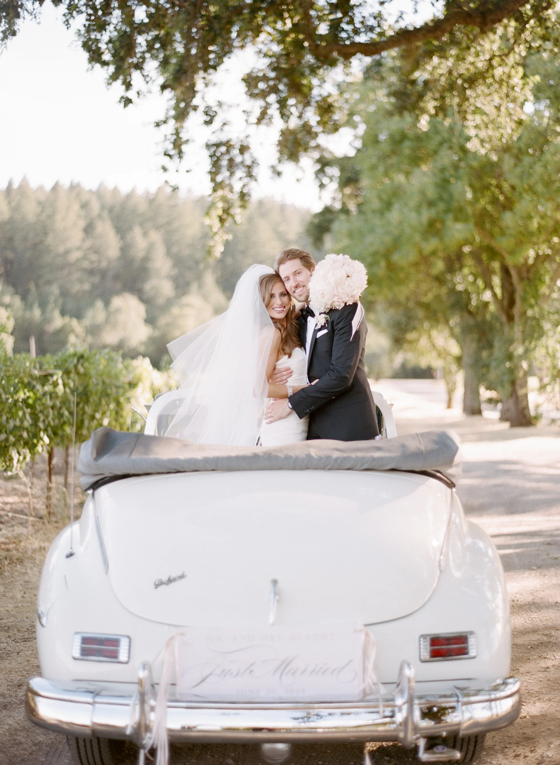 Lauren &amp; Michael embrace, standing in a vintage white car in a winery during a couple shoot with Sylvie Gil