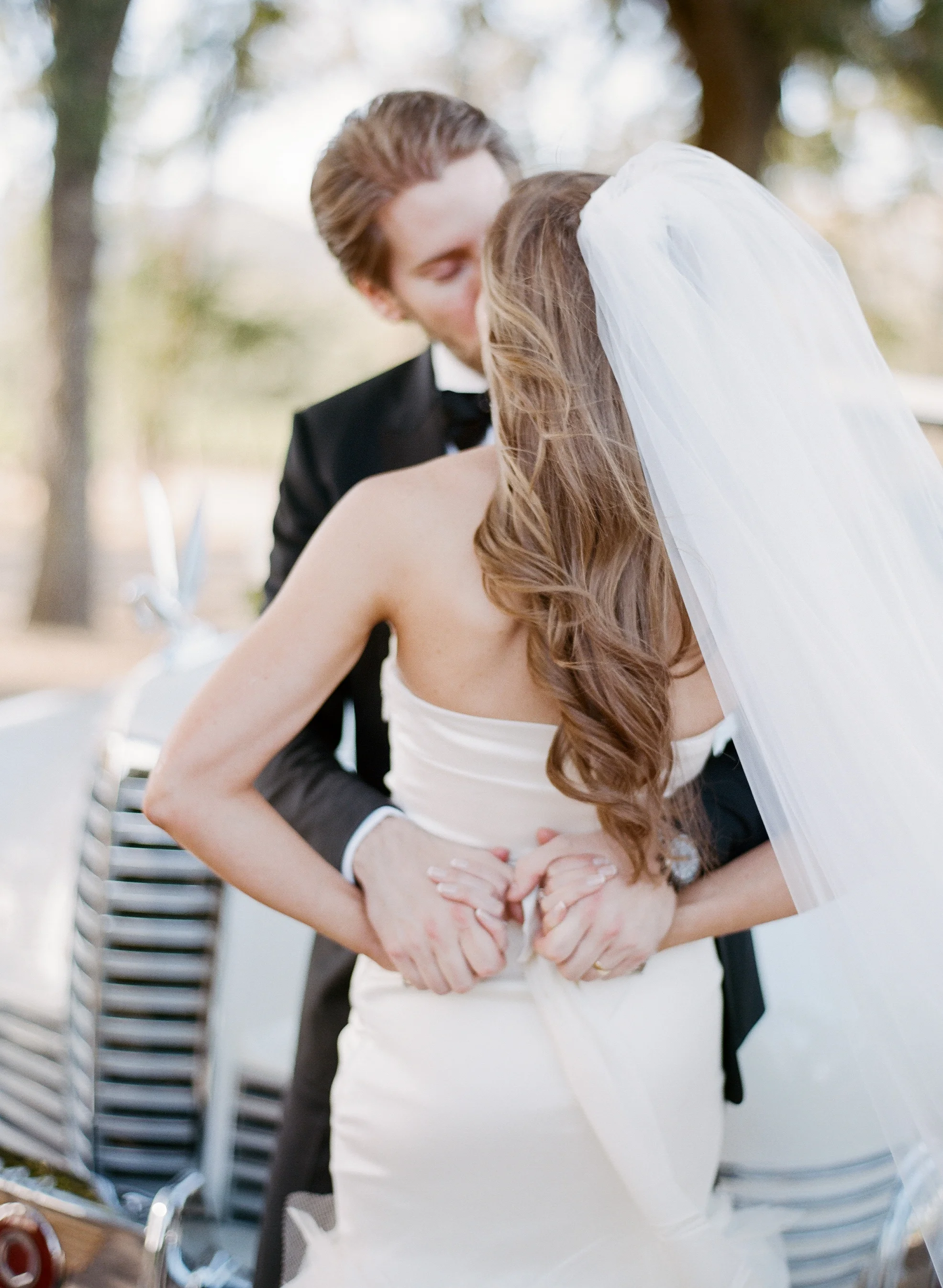 The couple shares a kiss in front of a vintage white car in a winery during a couple shoot with Sylvie Gil