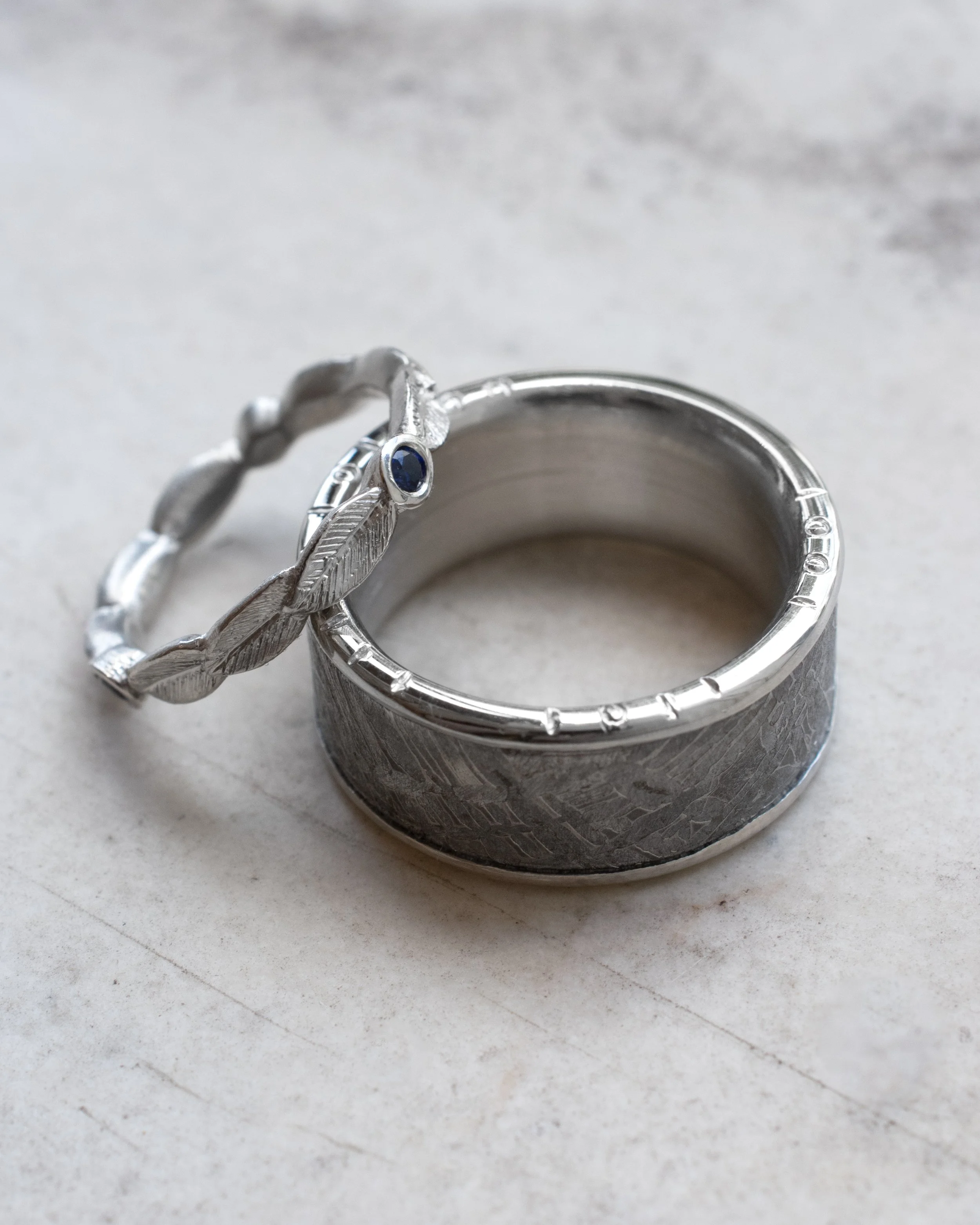 Two platinum wedding rings, one with textured surface and one with a feather design with a blue sapphire, placed on a light-colored surface.