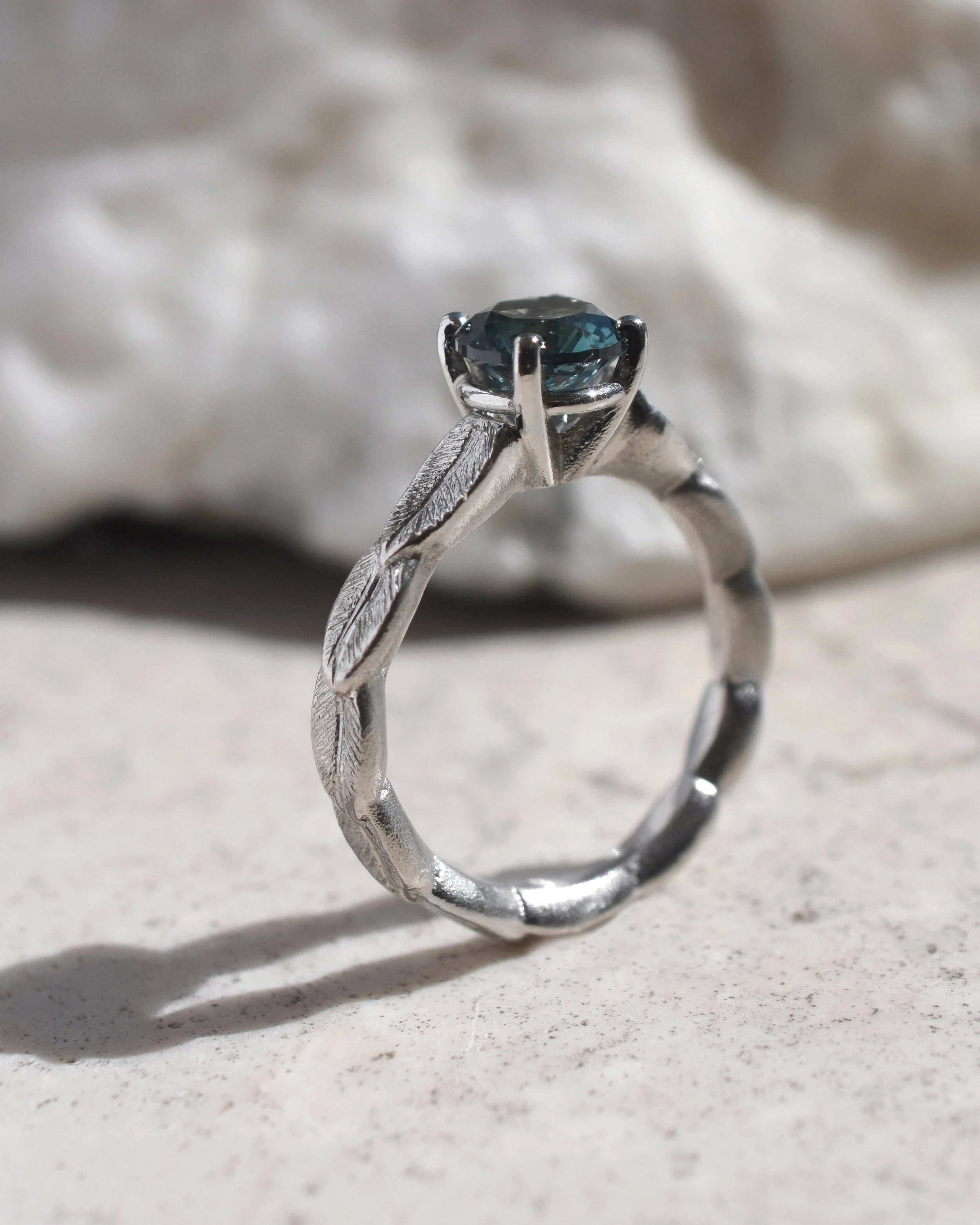 A platinum ring with a nature inspired design, featuring a American mined Montana sapphire in a prong setting, resting on a light-colored surface with a decorative white background.