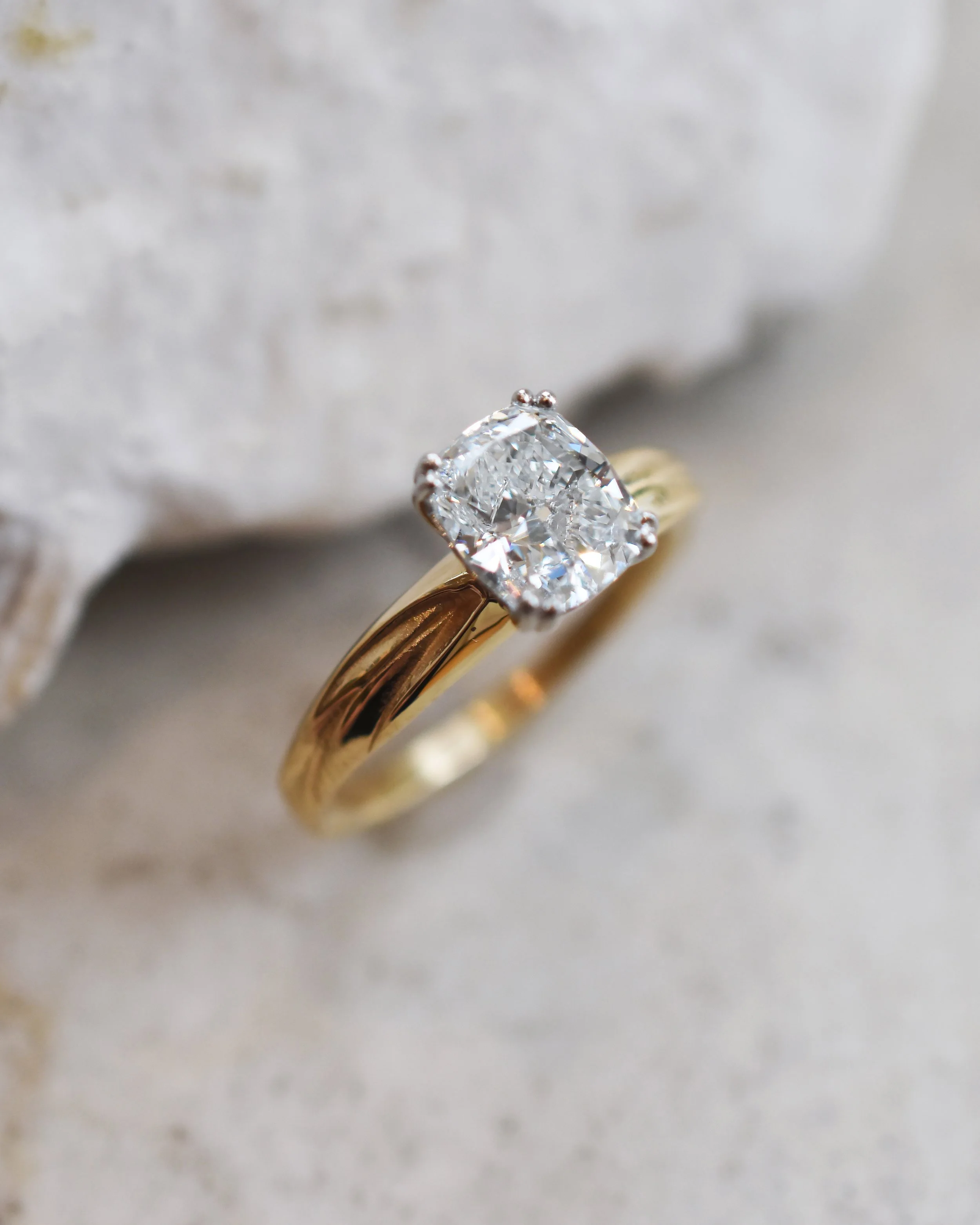 Close-up of a gold engagement ring with a prominent diamond in a prong setting on a light, textured surface.