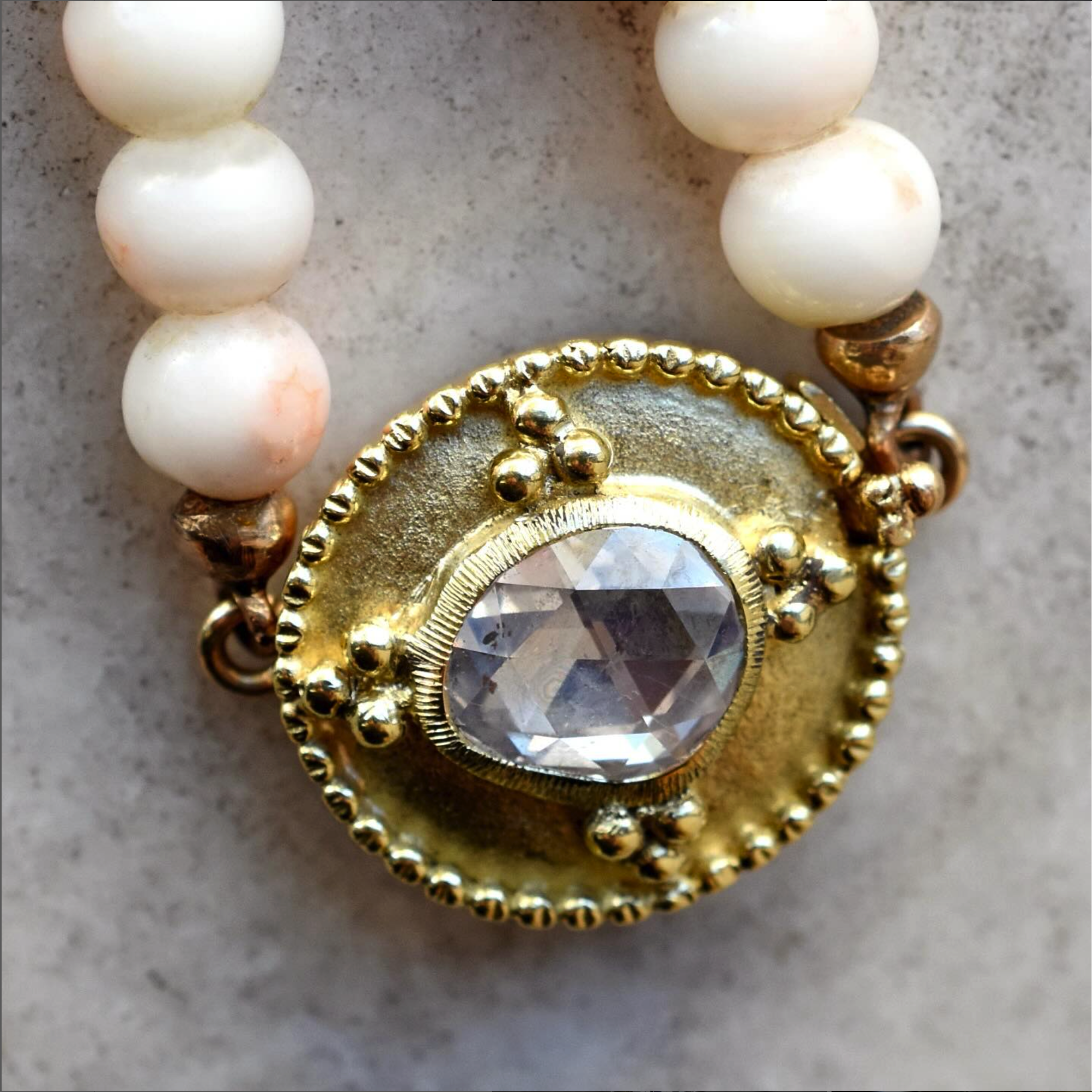 Close-up of a vintage-style jewelry piece featuring a central diamond-like gemstone, surrounded by gold and beaded detailing, attached to a string of white pearls.