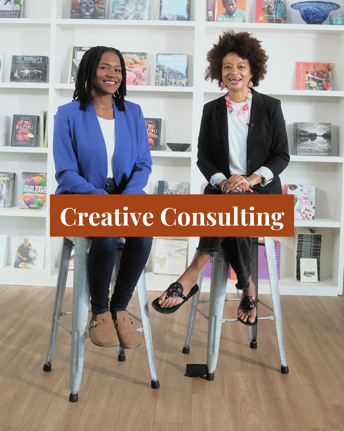 Two women sitting on high stools in front of a bookshelf filled with books, with a sign that reads 'Creative Consulting' in front of them, suggesting a professional or creative setting.