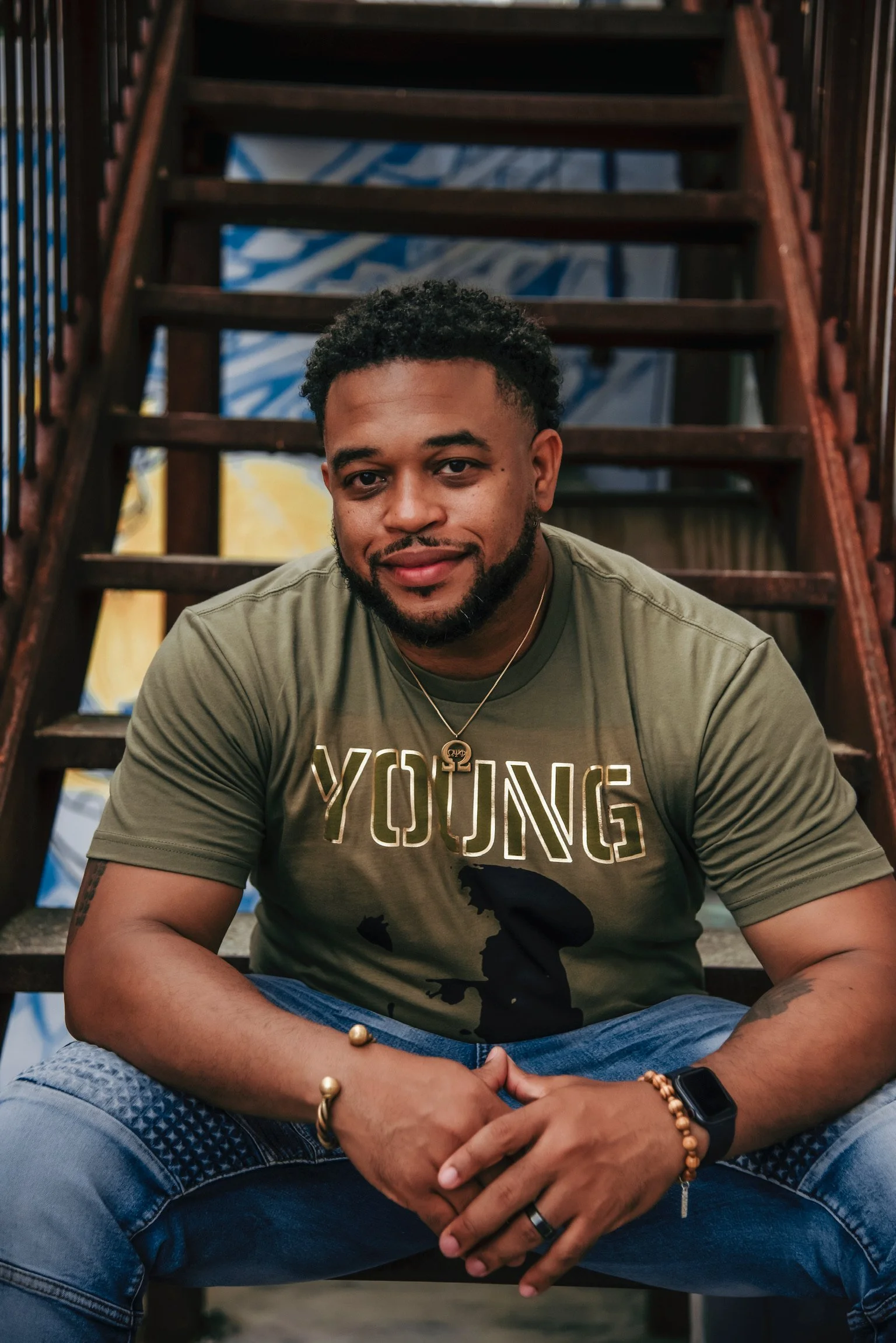 A young man with short curly hair and a beard sitting on outdoor stairs, wearing a green t-shirt with the word 'YOUNG' and a map silhouette, blue jeans, wristbands, and jewelry, smiling at the camera.