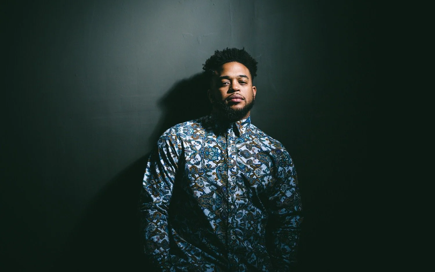A man with curly hair and a beard wearing a patterned shirt, standing against a dark wall, looking at the camera.