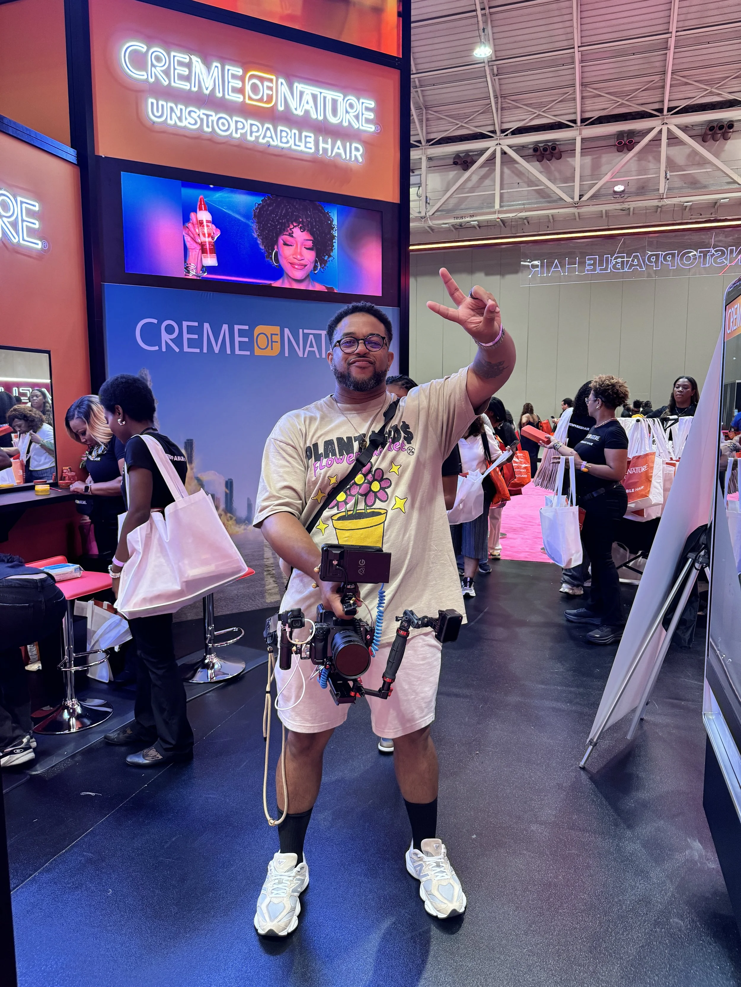 A man with glasses, a beard, and tattoos makes a peace sign with his right hand while standing in a convention center. He is wearing a beige t-shirt with colorful graphics, light shorts, and chunky white sneakers. There are people and booths in the background, and large signs promoting "Creme of Nature" and "Unstoppable Hair." The ceiling of the convention hall is visible with white beams and lighting.