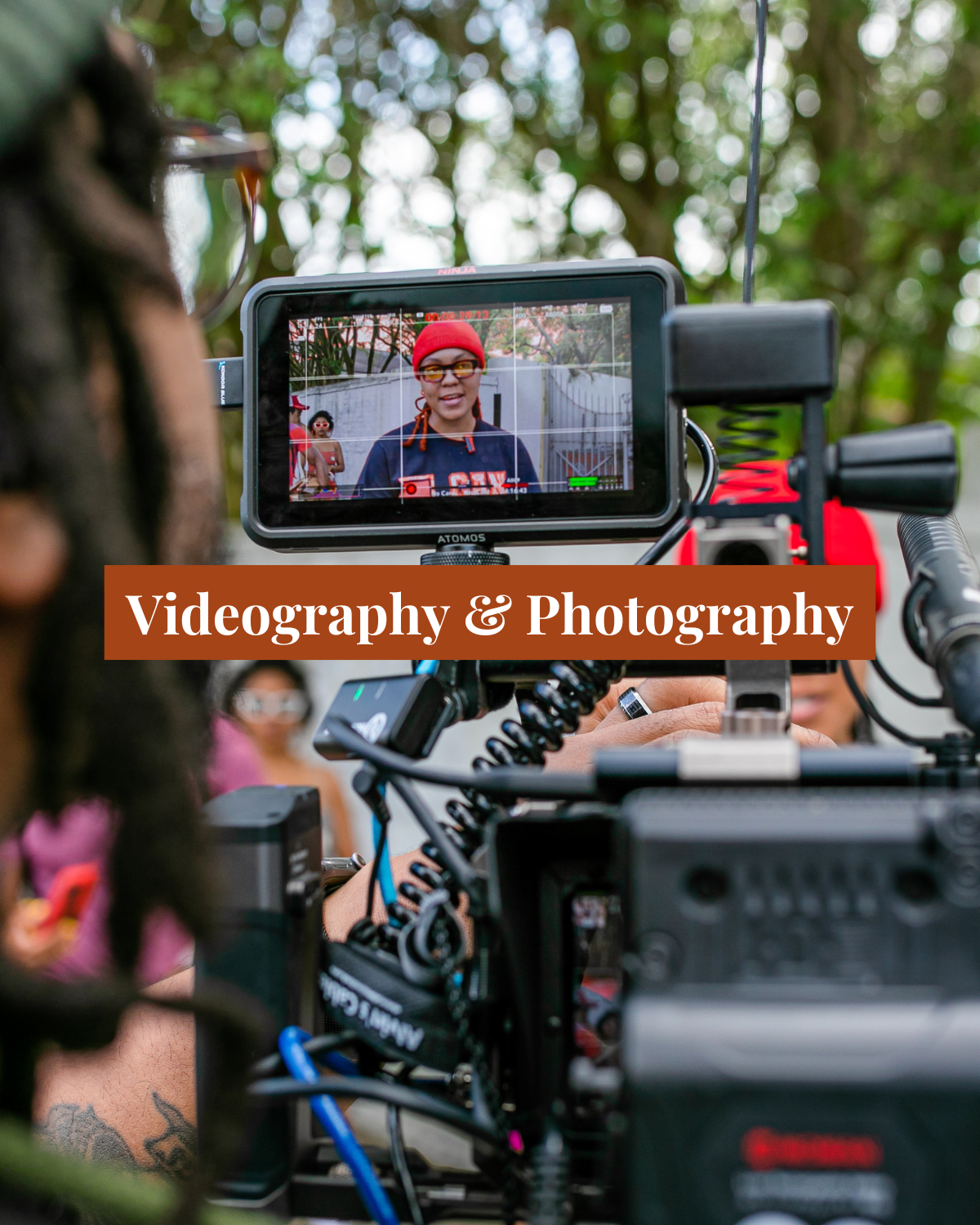 A video camera filming a woman with red hair, glasses, and a red beanie, outdoors with a green background. The camera's screen shows the woman smiling and speaking, with others visible in the background. Overlaid text reads 'Videography & Photography'.