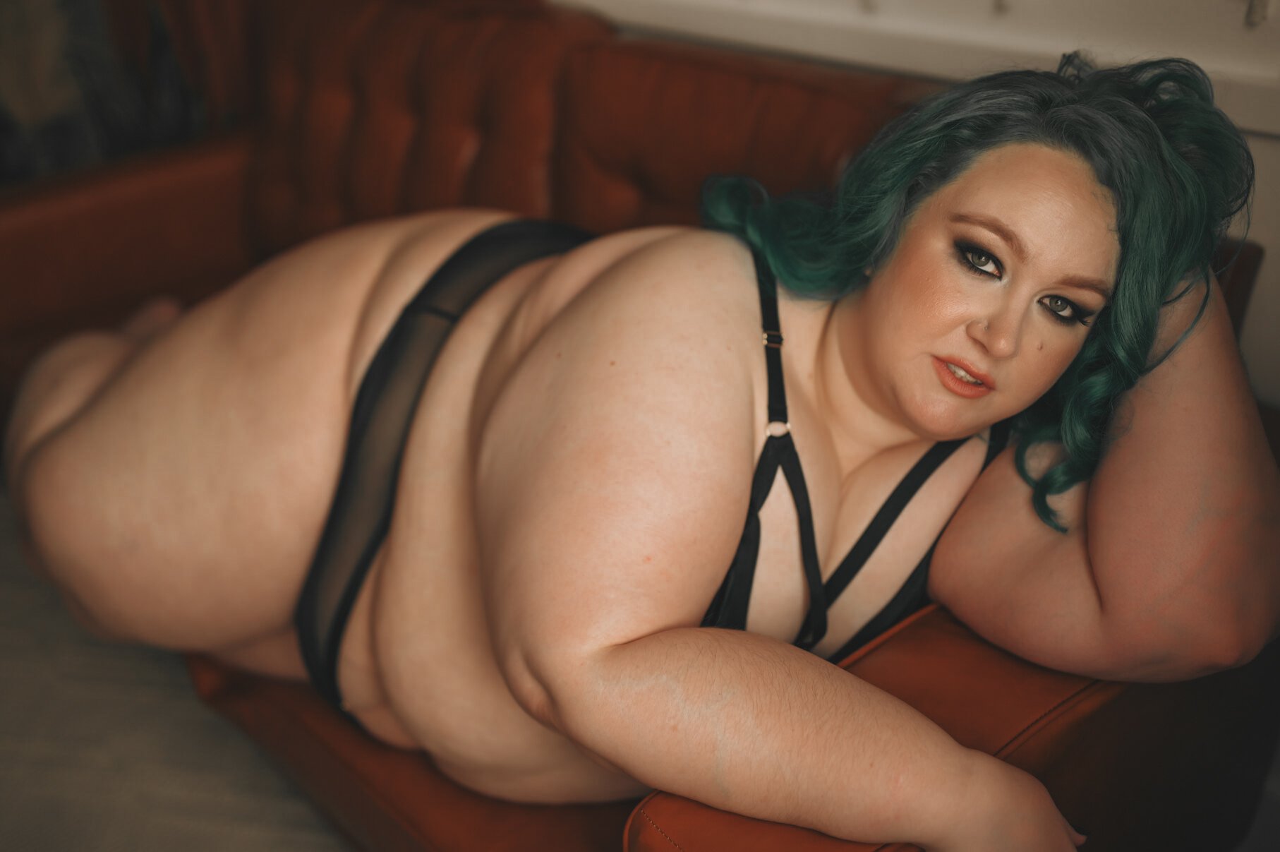 A plus-size woman with teal hair and smoky eye makeup lounges on a brown leather sofa in black lingerie, gazing confidently at the camera—capturing the empowering essence of boudoir photos.