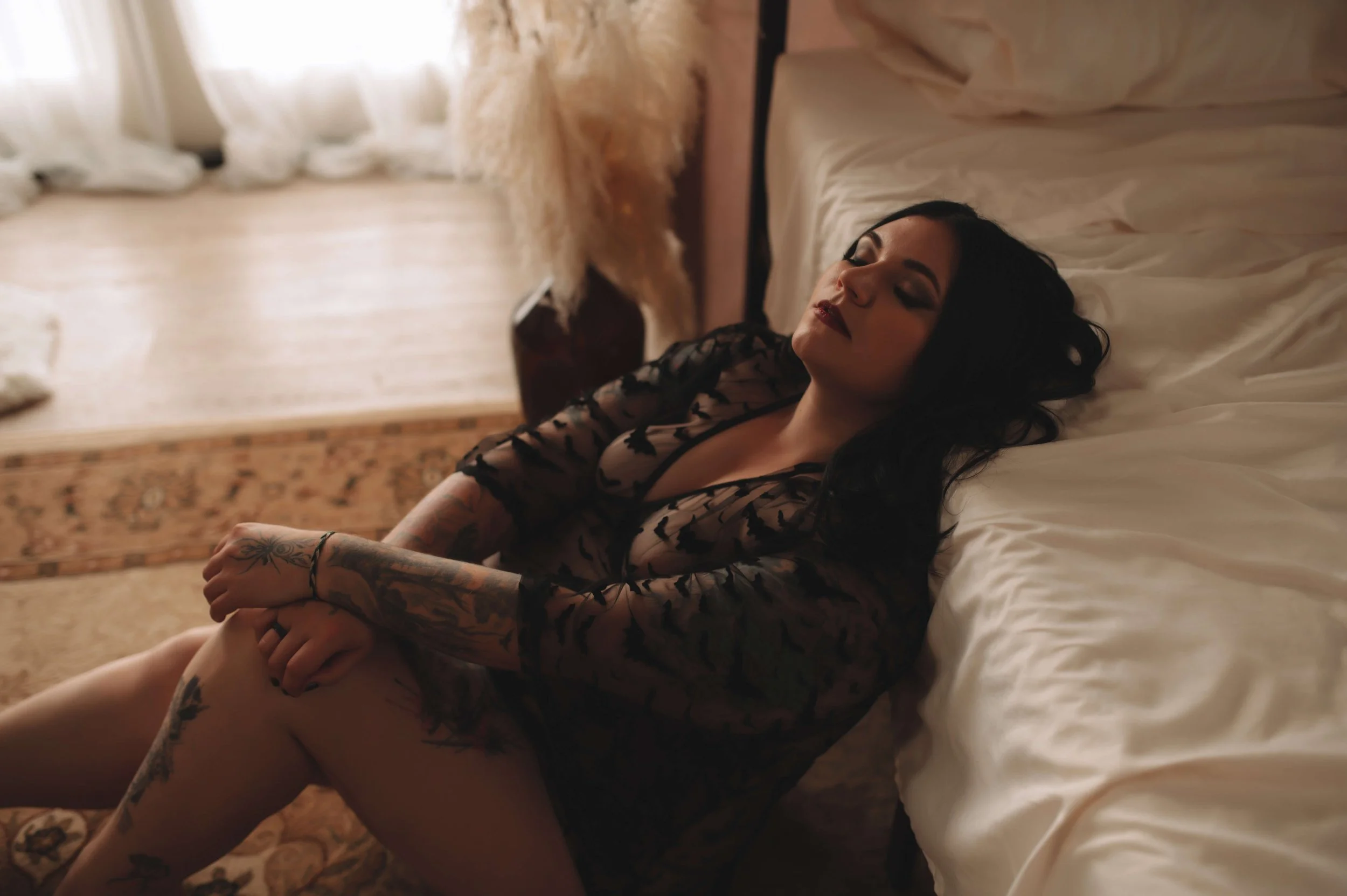 A woman with black hair and tattoos wearing a black lace dress, sitting on the floor with her back against a bed, in a softly lit room.