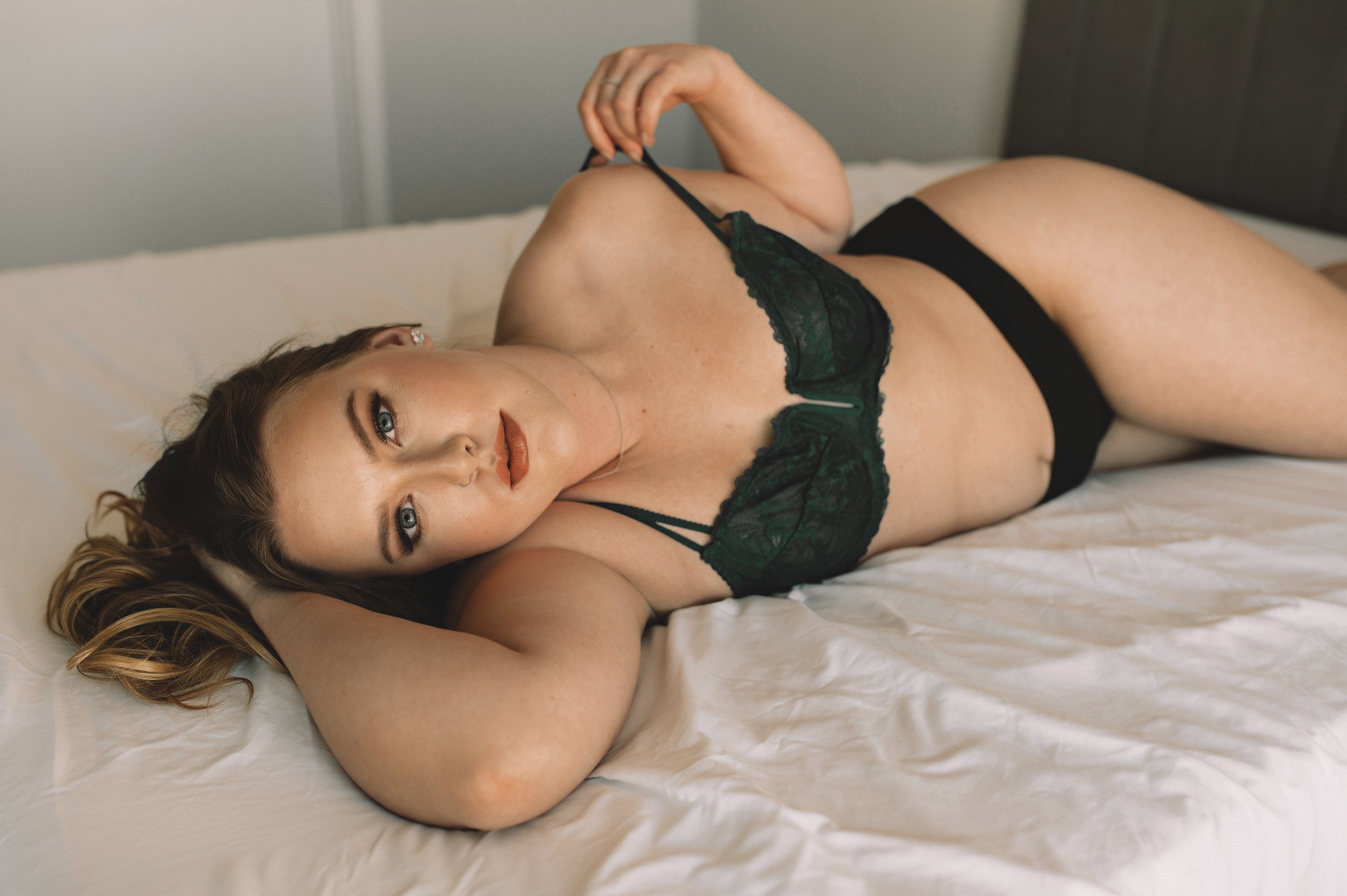 A woman in black lingerie lies on a bed with white sheets, resting her head on one arm and looking at the camera while gently holding her bra strap—capturing the intimate allure of boudoir photos.