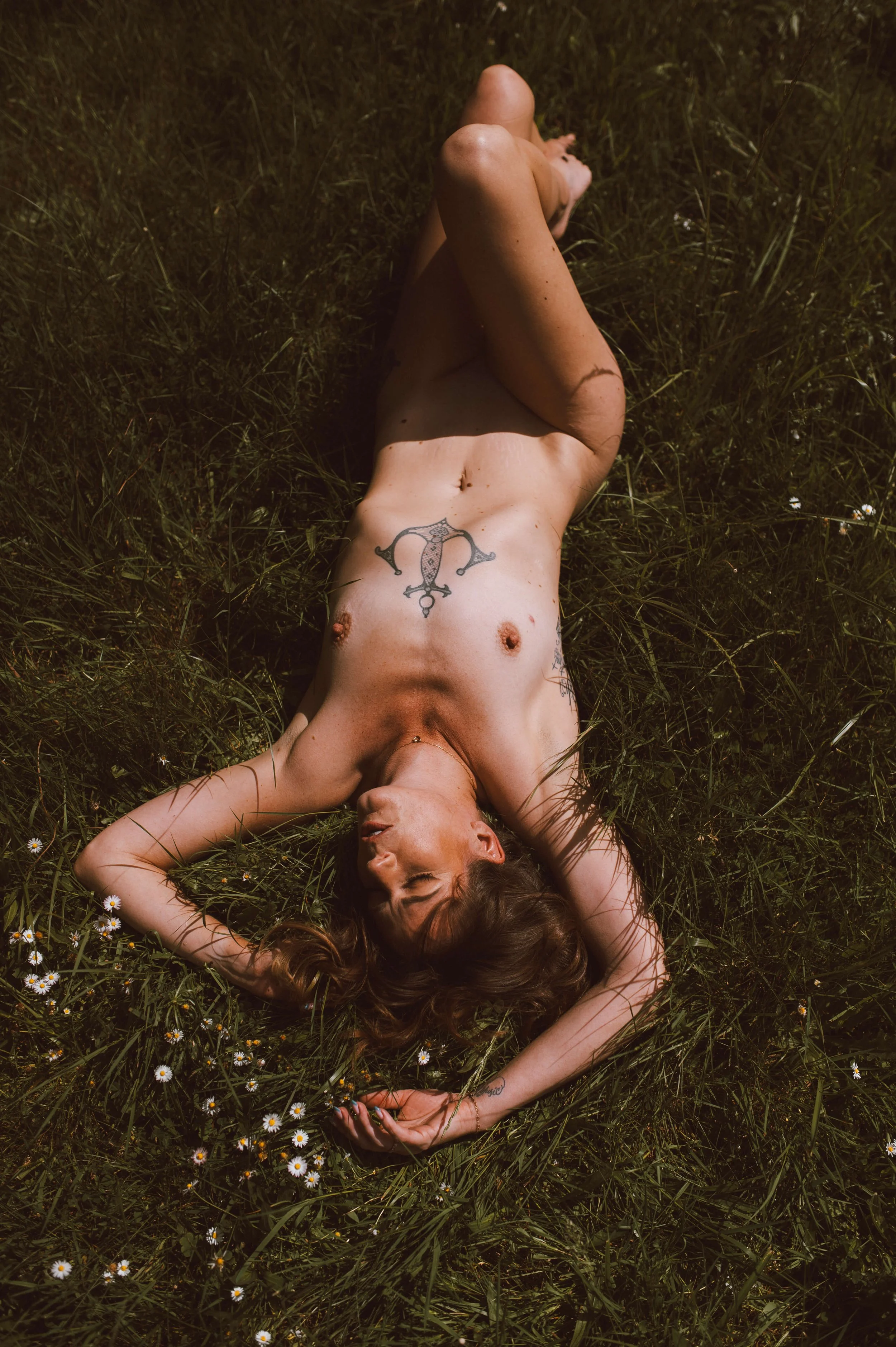 A nude woman with long, wavy hair is lying on her back in a grassy field, surrounded by small white flowers, with one leg crossed over the other and her eyes closed.