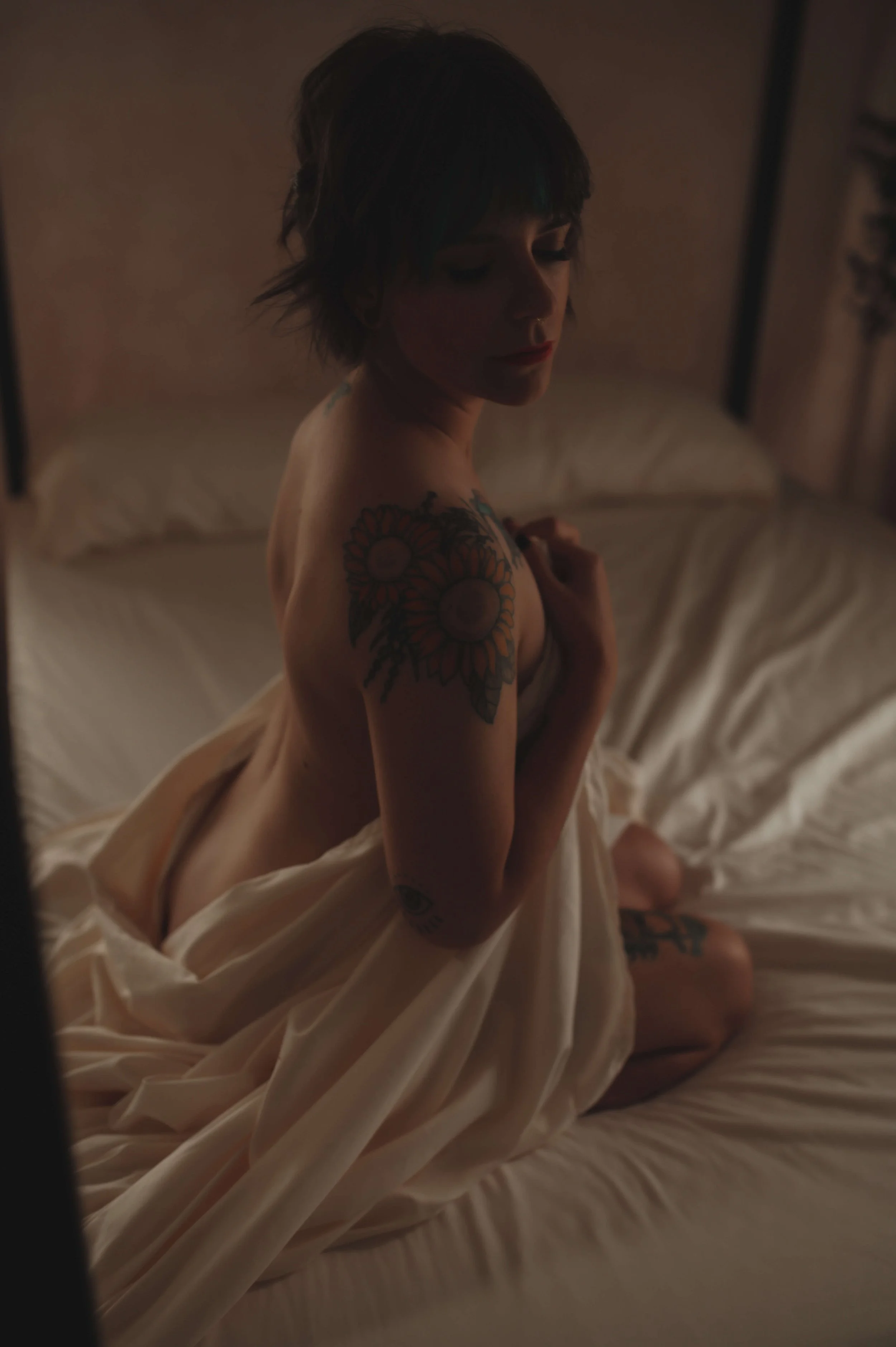 A woman with short dark hair and tattoos is kneeling on a bed, holding a blanket across her chest, in a dimly lit room.