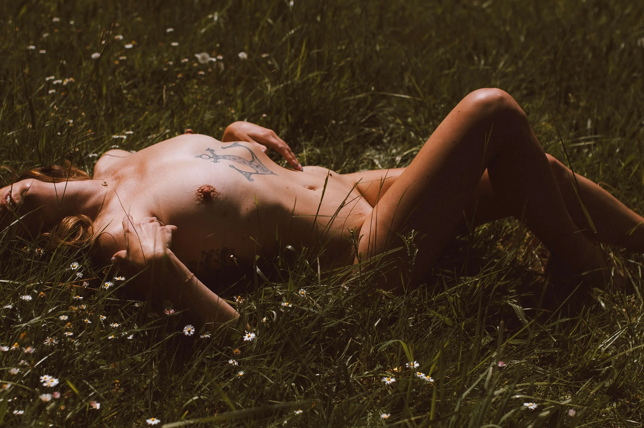 A topless woman with tattoos lying on her back in a grassy field with flowers, sunlight illuminating her skin.
