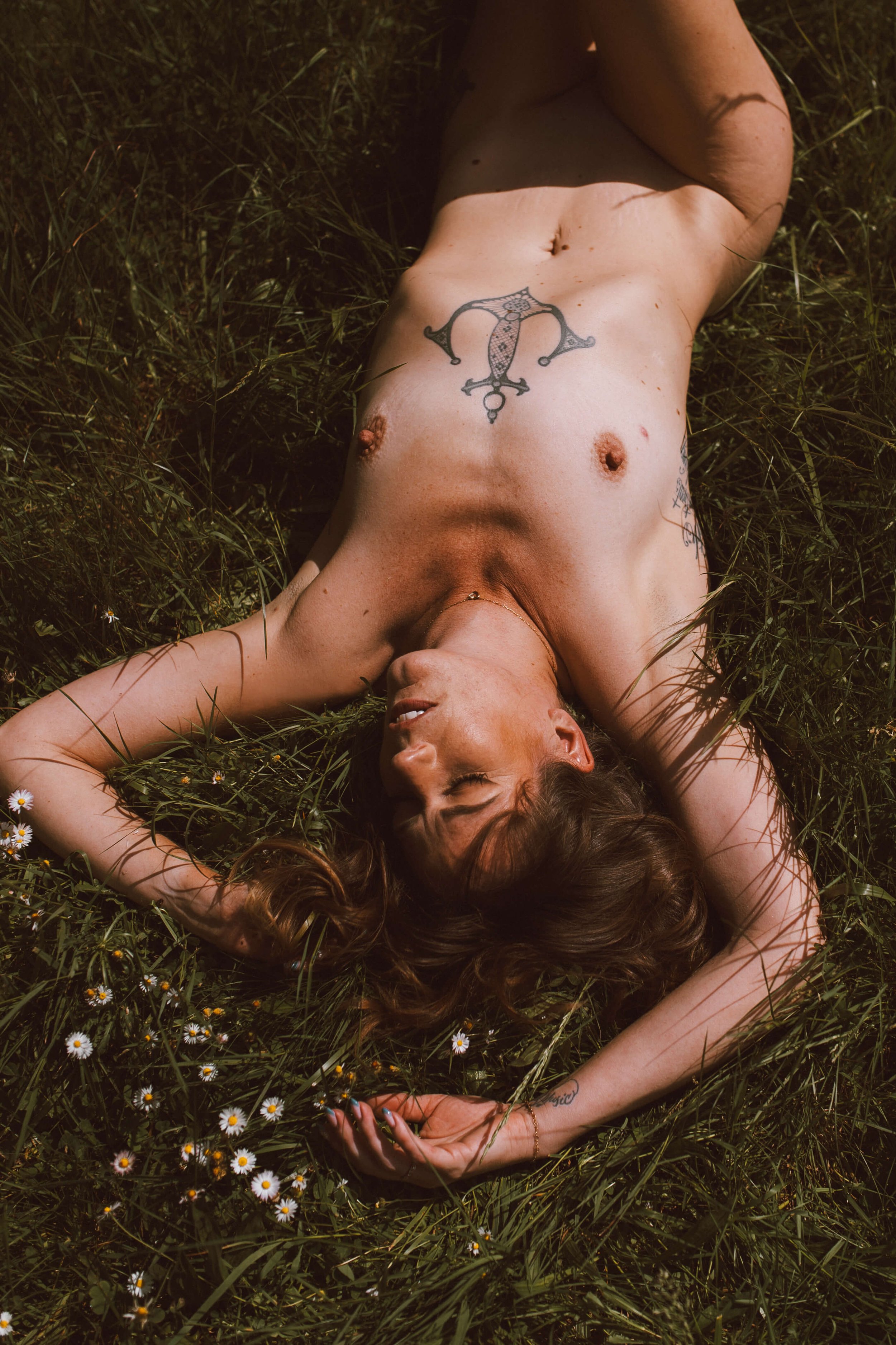 A woman with long brown hair, lying on her back in a grassy field with small white flowers, with her hands over her head and her eyes closed.