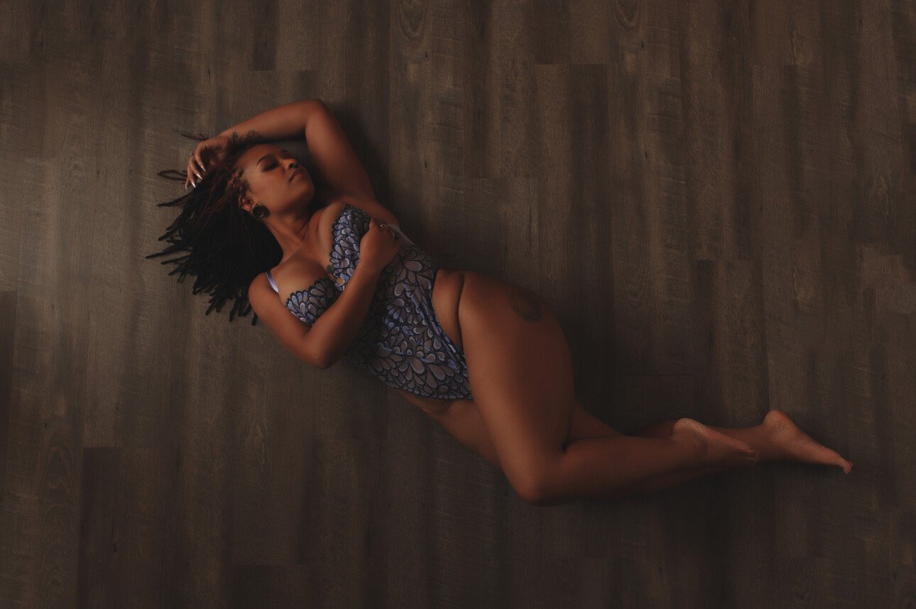 A woman with dark braided hair lies on a wooden floor, wearing a blue patterned one-piece swimsuit. Posed as if for boudoir photos, she rests one arm across her chest and the other above her head, legs slightly bent.