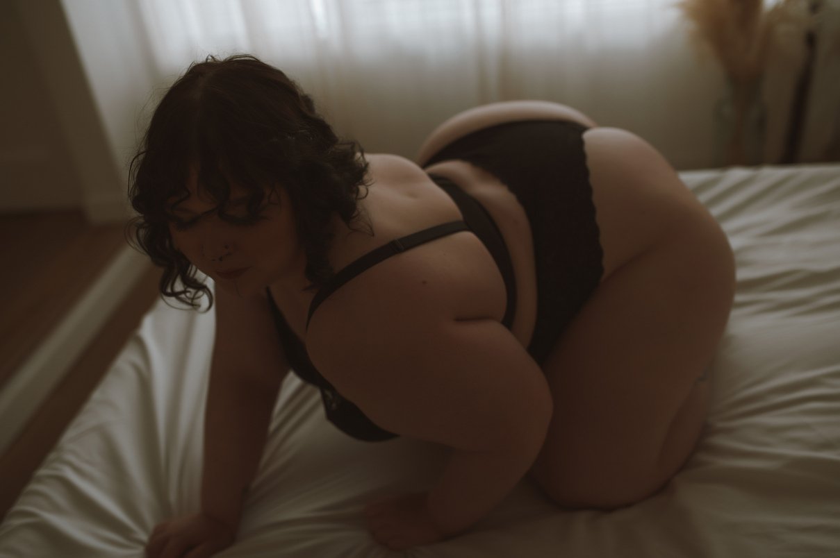 A woman with curly dark hair, dressed in black lingerie, poses on a bed with white sheets in a softly lit room. Light filters through the window, capturing the intimate vibe of boudoir photos with personalized attention to detail.