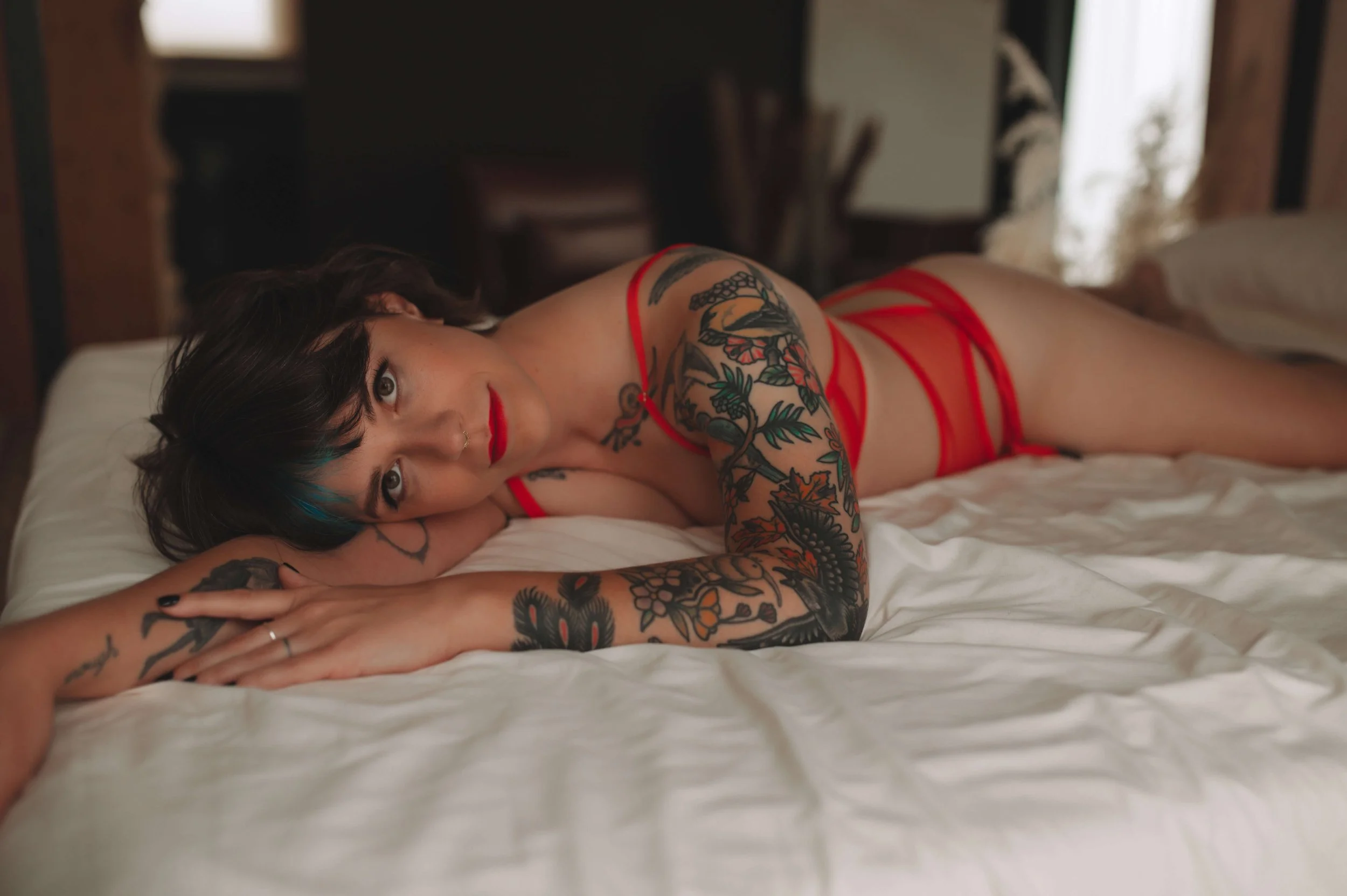Woman with black hair and tattoos lying on a bed wearing red lingerie, looking at the camera.