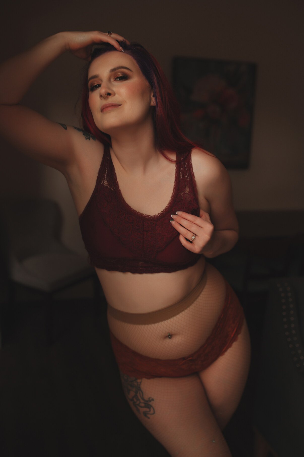 A woman with long reddish hair poses confidently indoors for boudoir photos, wearing a burgundy lace bralette and matching lingerie. She has a hand on her head, smiling softly, and a tattoo is visible on her upper thigh.